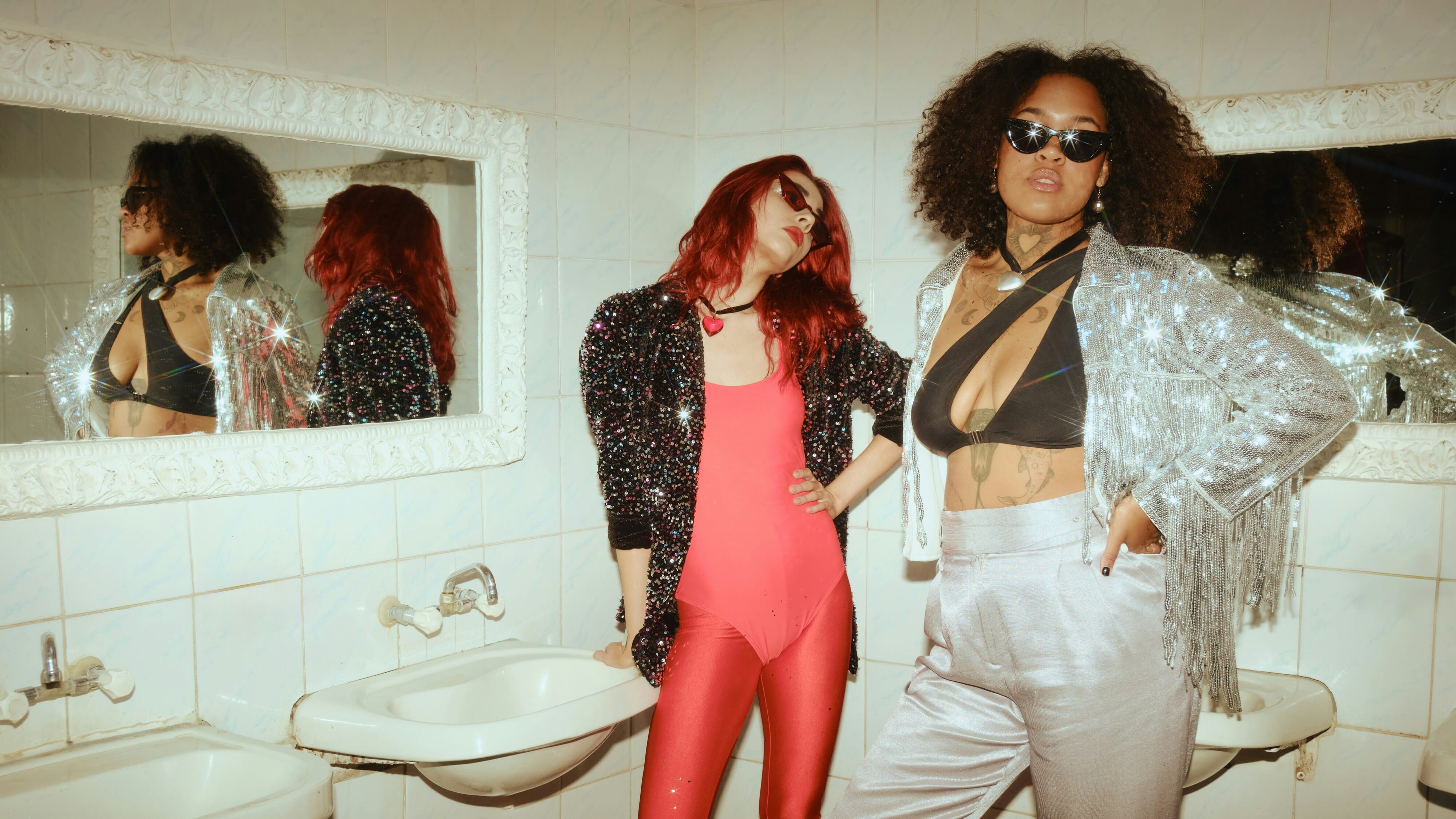 Zwei Frauen vor Wäschbäcken in einem glitzernden Party Outfit. 