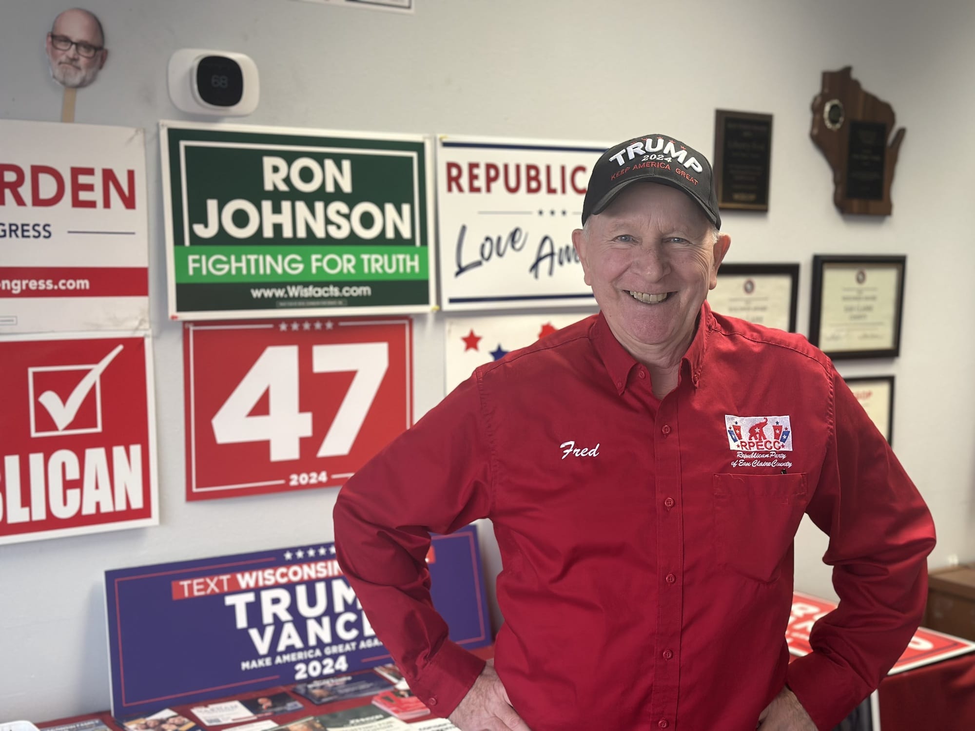 Man sieht einen älteren Mann in rotem Hemd vor politischen Schildern.