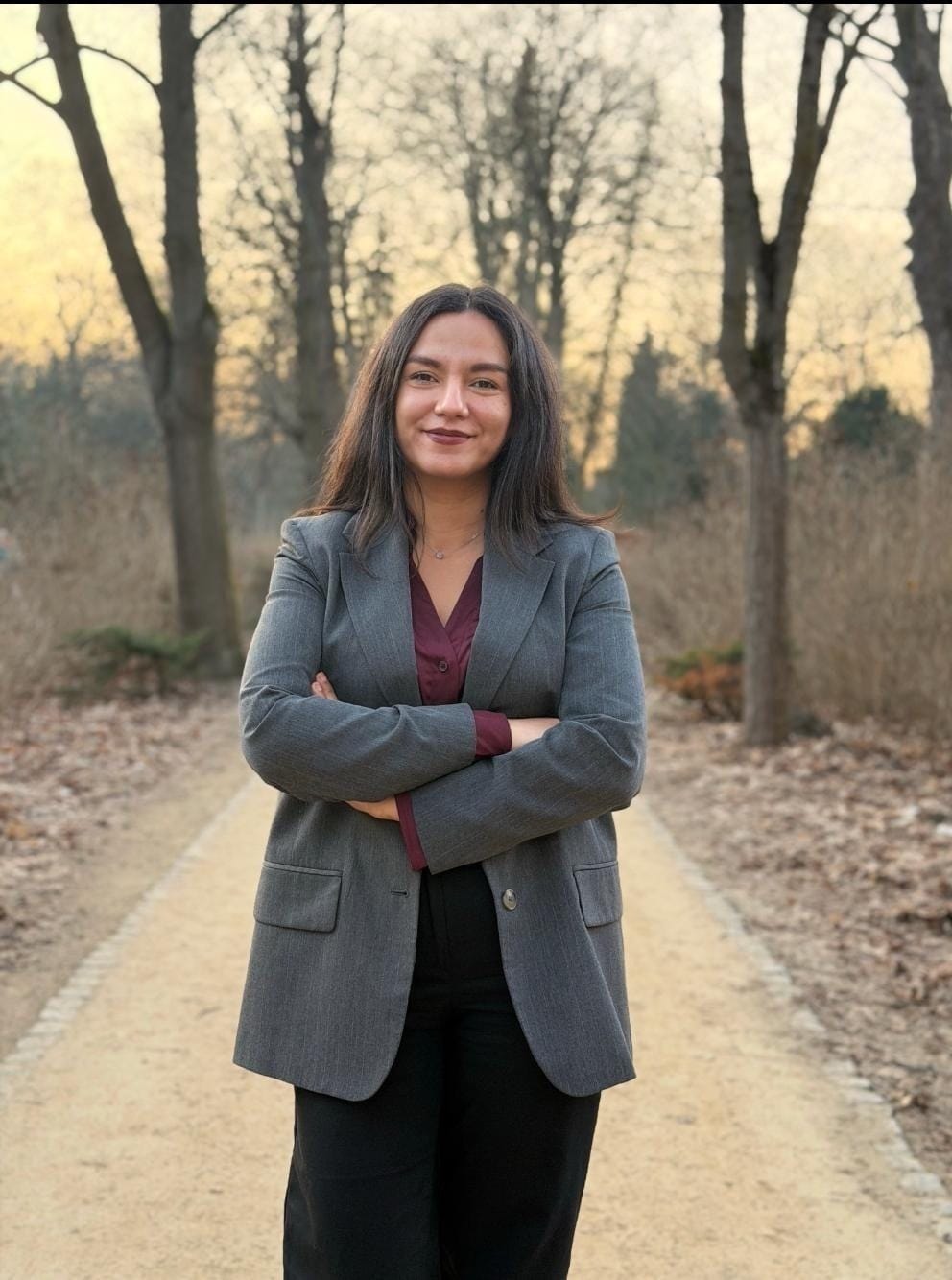 Basma steht in einem Park und lächelt mit verschränkten Armen in die Kamera.