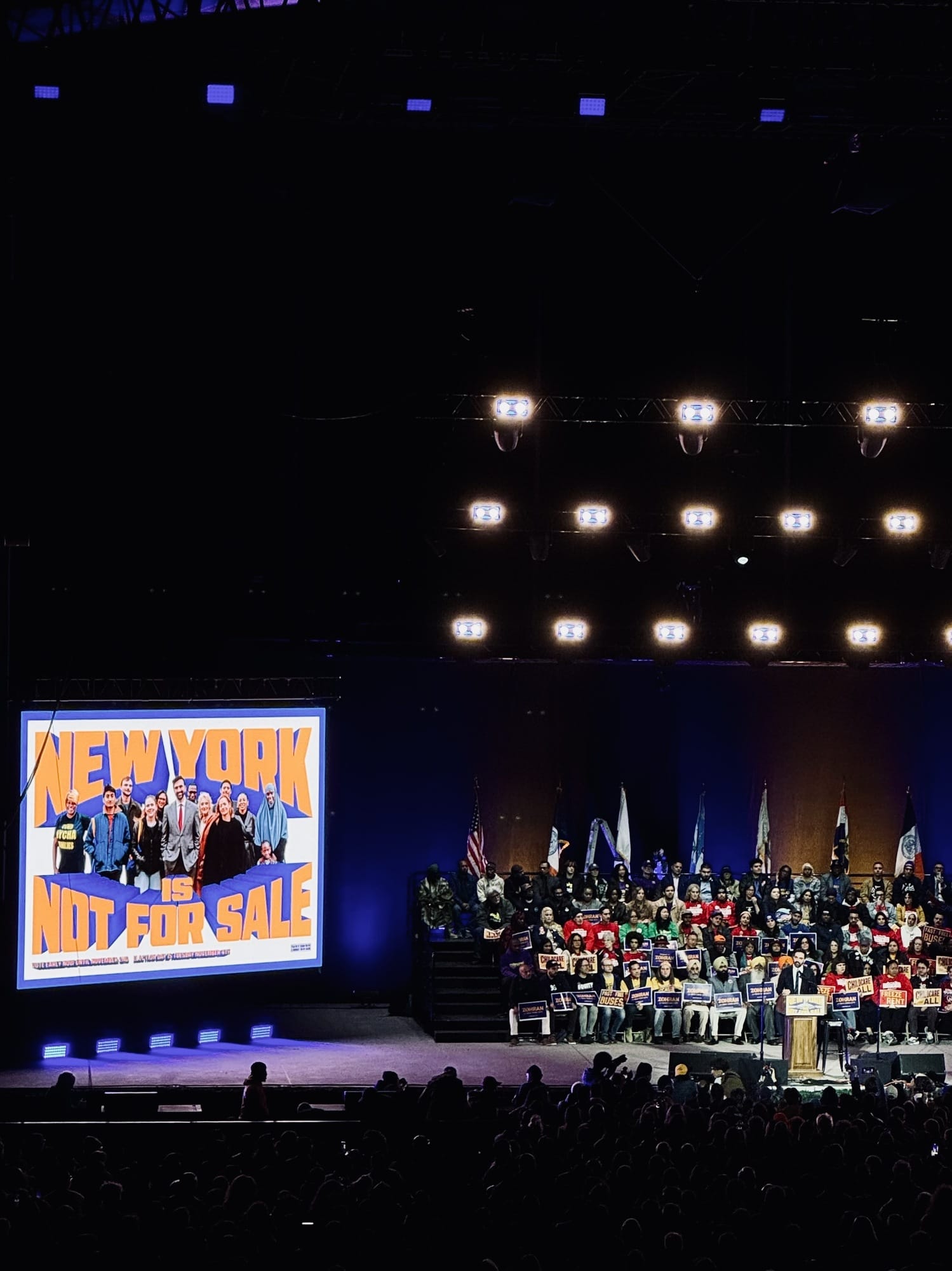 Man sieht eine große Bühne, links ein Bildschirm auf dem steht "New York is not for sale", rechts eine Menschenmenge auf der Bühne.