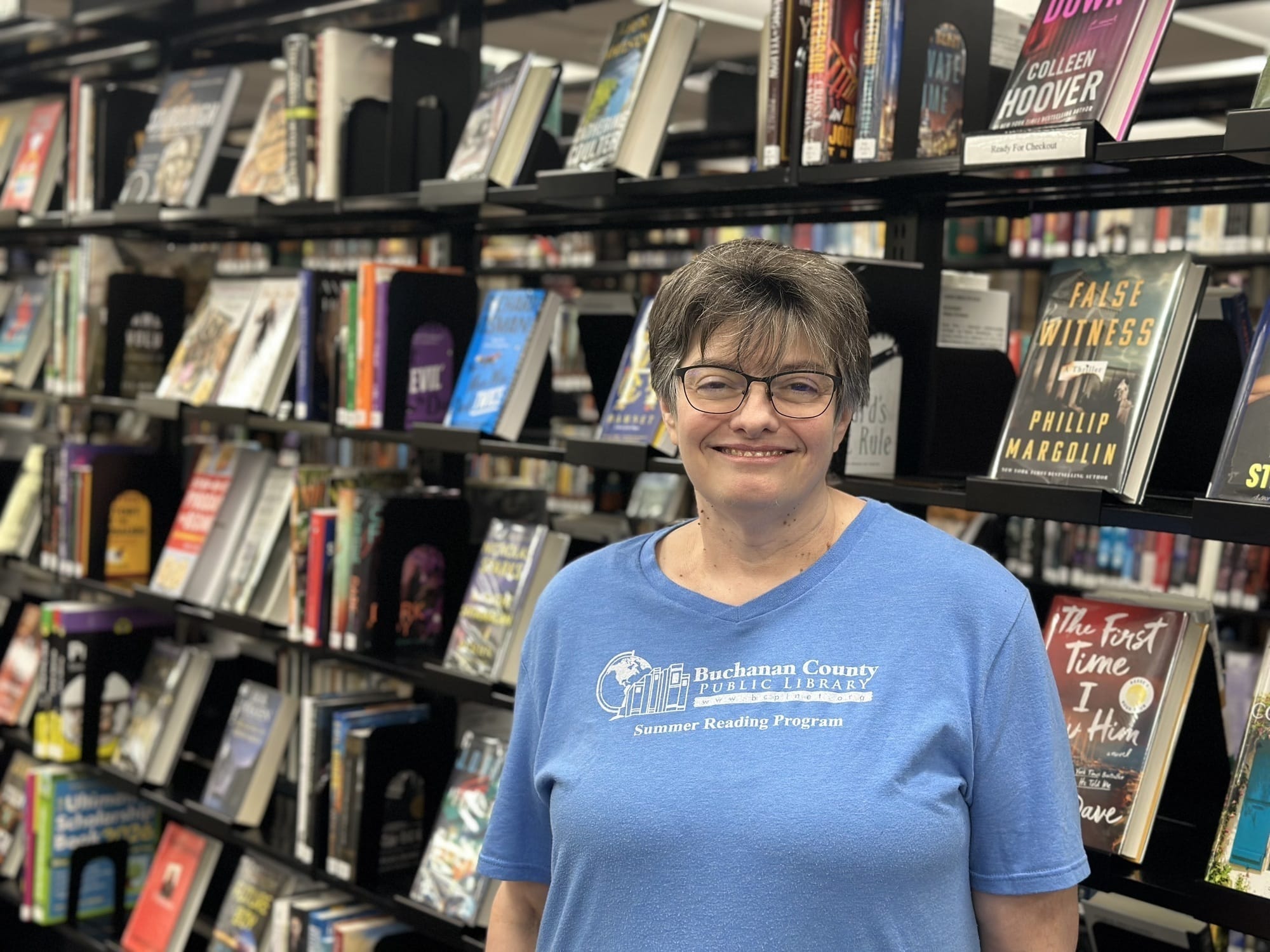 eine Frau in blauem T-Shirt steht vor einem Bücherregal und lächelt.