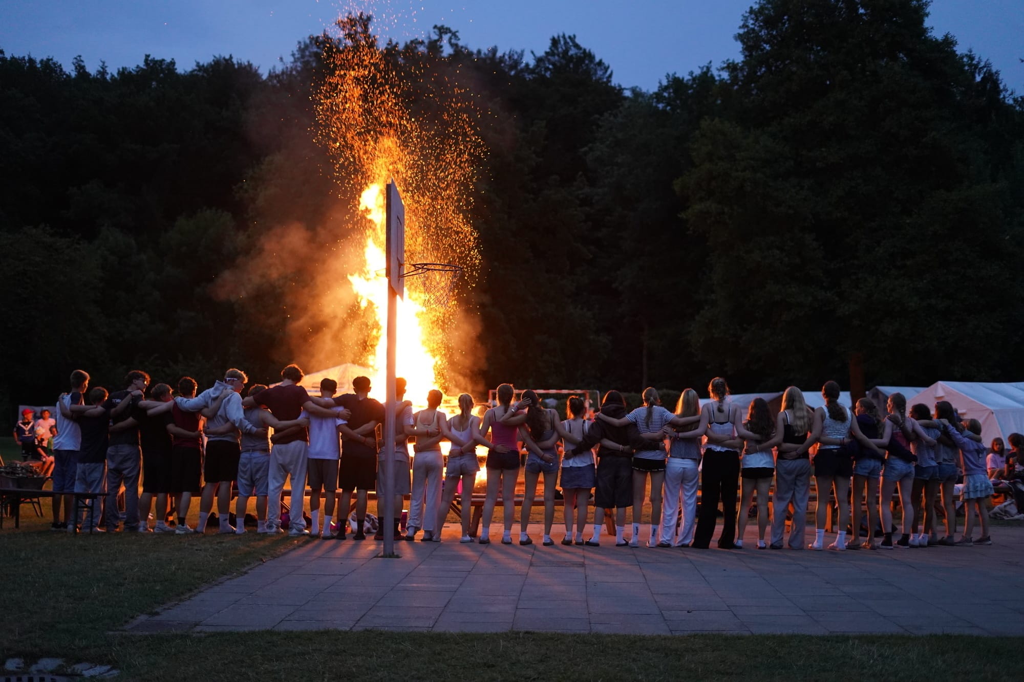 Eine Reihe von Kindern und Jugendlichen, die Arm und Arm um ein Lagerfeuer herum stehen.