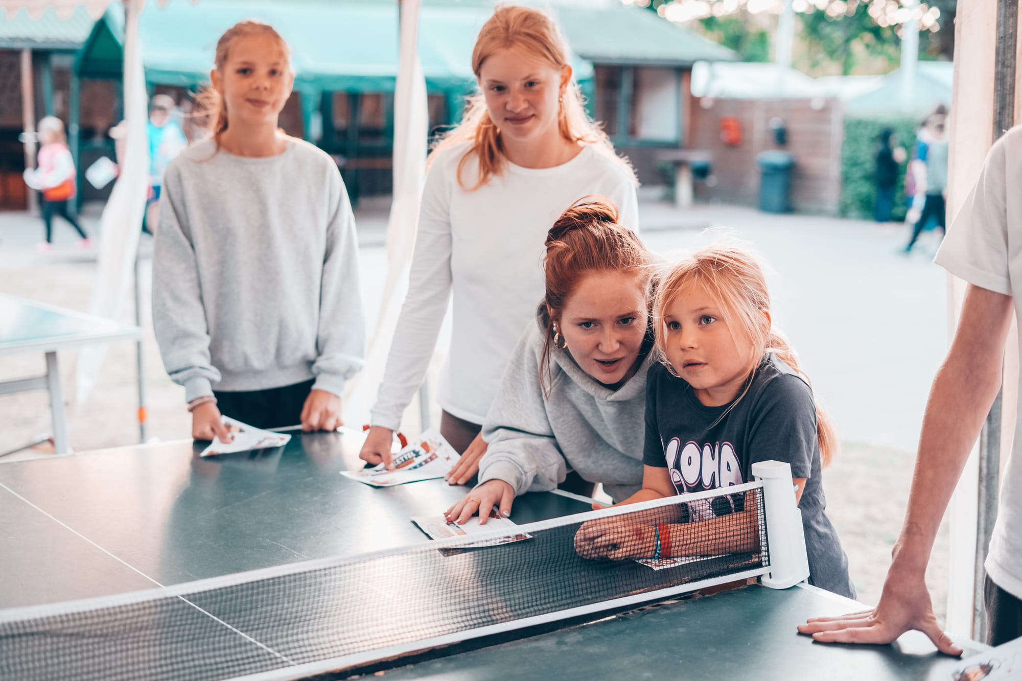 Ein siebenjähriges Mädchen steht an einer Tischtennisplatte. Eine 15-jährige bückt sich zu ihr herunter und spricht mit ihr. 
