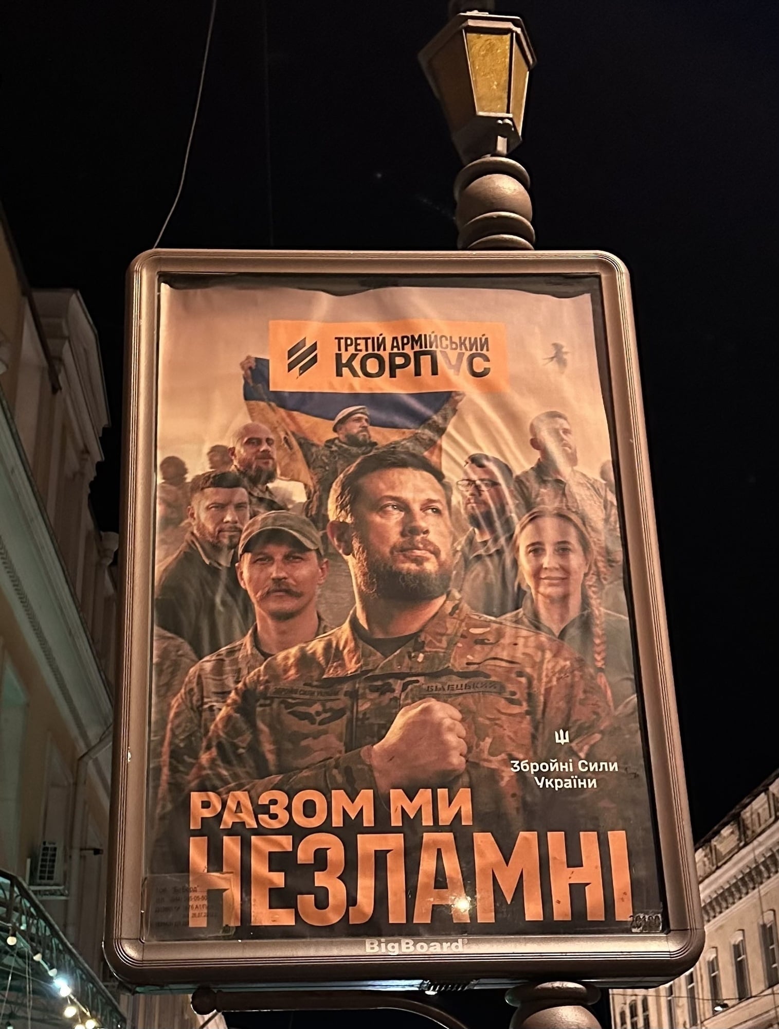 Foto eines Plakats an einem Laternenpfahl. Man sieht mehrere lächelnde Menschen und ukrainische Schrift.