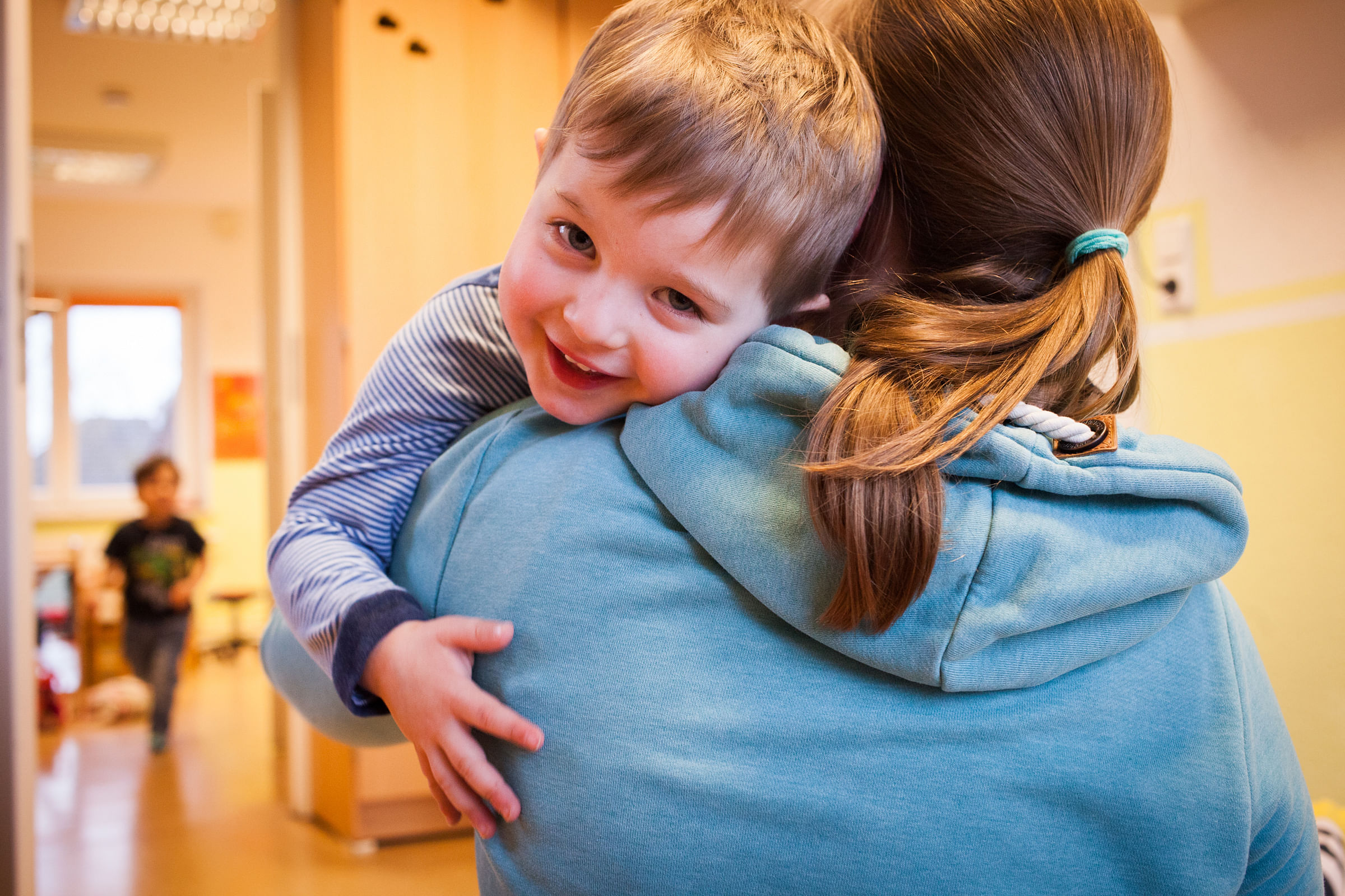 Man sieht das Gesicht eines Jungen, der von der Erzieherin getragen wird. Der Junge lehnt sich an ihre Schulter an.