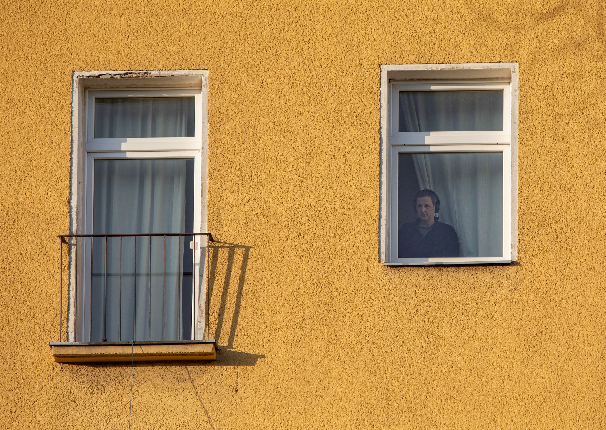 Wir sehen zwei Fenster, ein großes und ein kleines vor einer gelben Wand. Hinter dem kleinen Fenster steht ein Mann und schaut nach draußen.