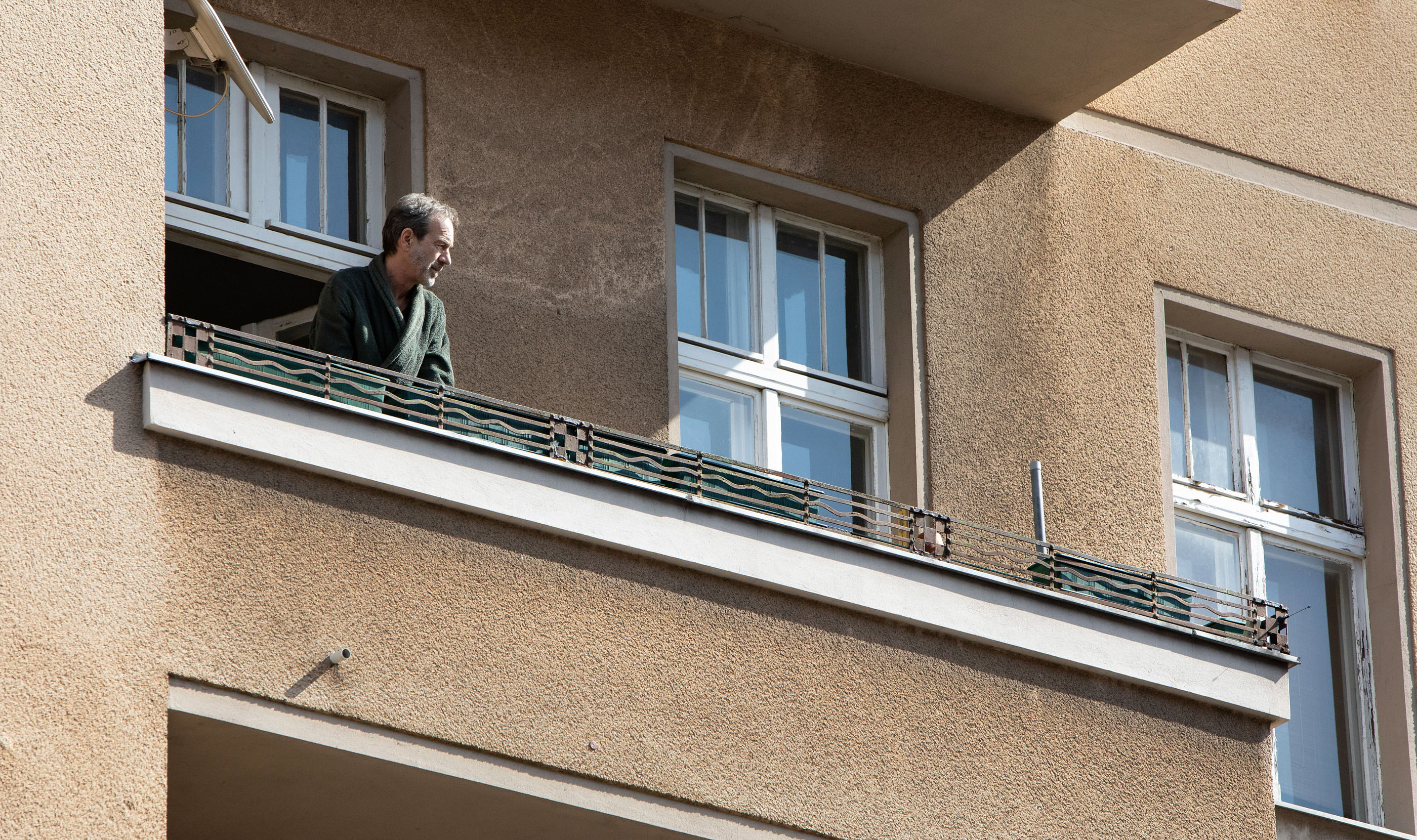 Ein Mann schaut von großen Balkon runter auf die Straße. Er trägt einen Bademantel.