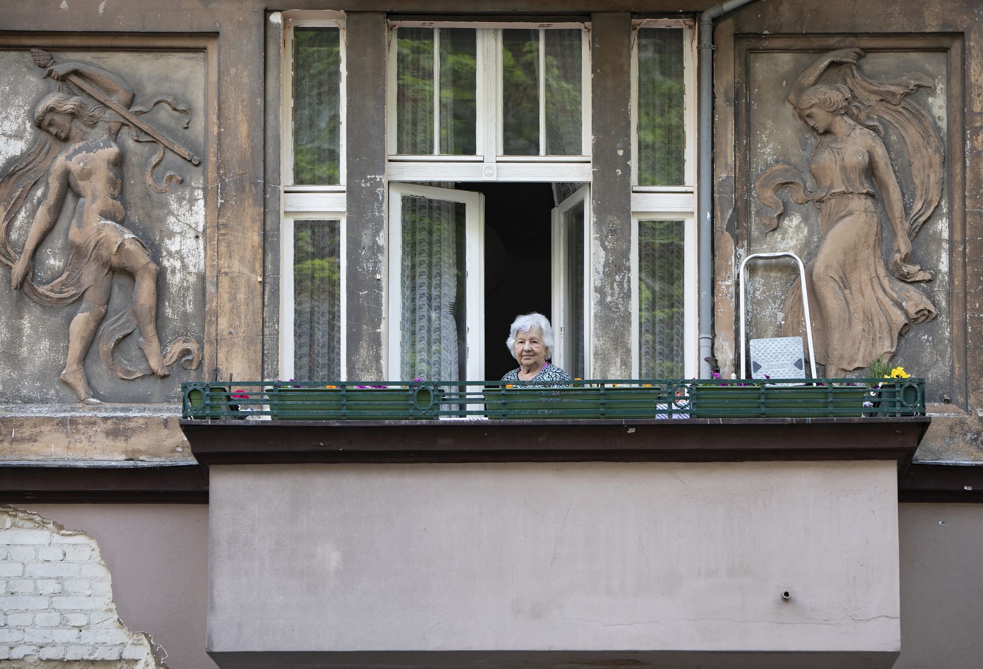 Eine Frau steht auf einem Balkon und sieht direkt in die Kamera. Hinter dem Balkon ist die Hauswand verziert mit Kunst.