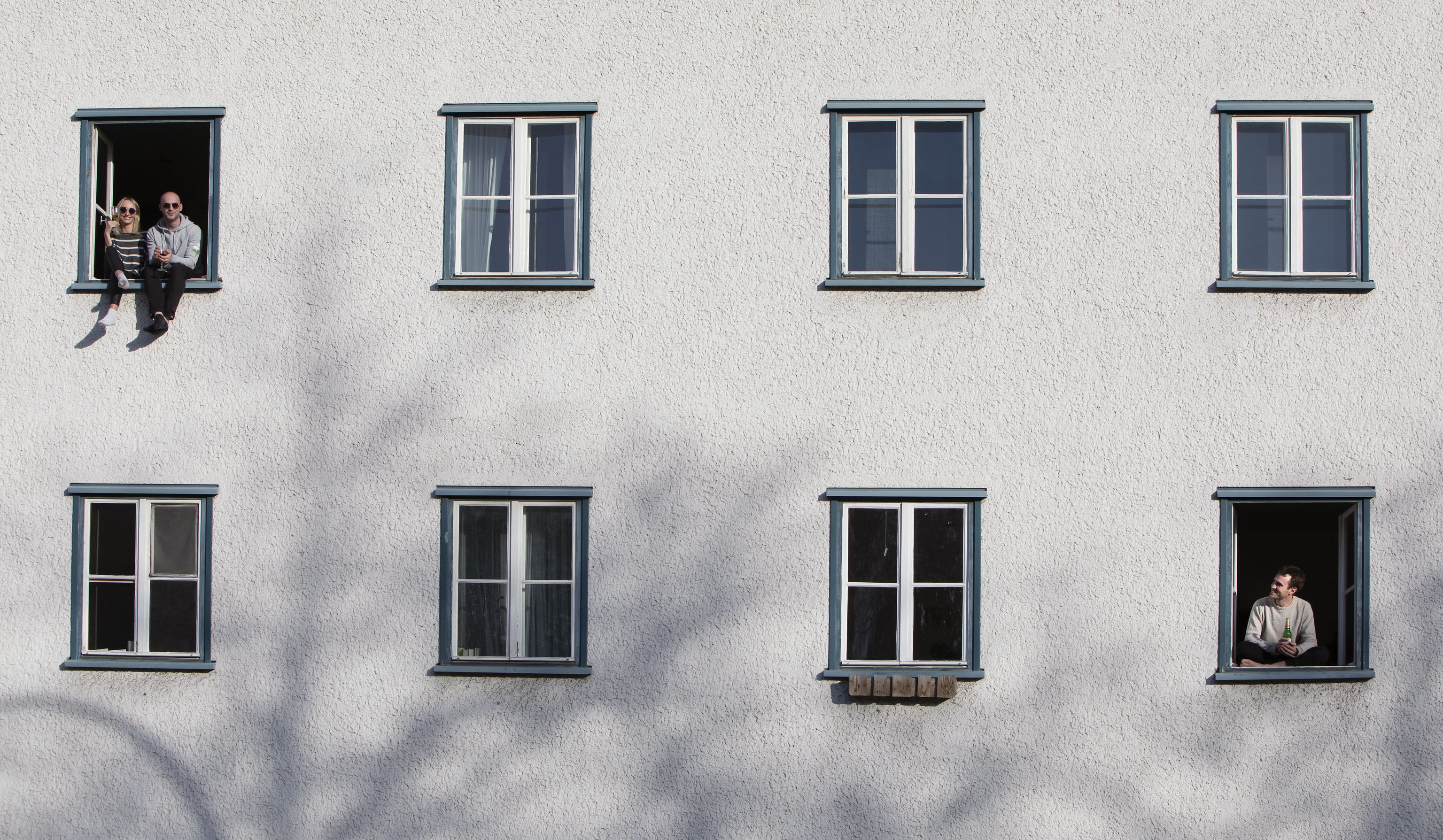 Eine Hauswand mit acht Fenstern. Links oben und rechts unten gucken Menschen Richtung Kamera.