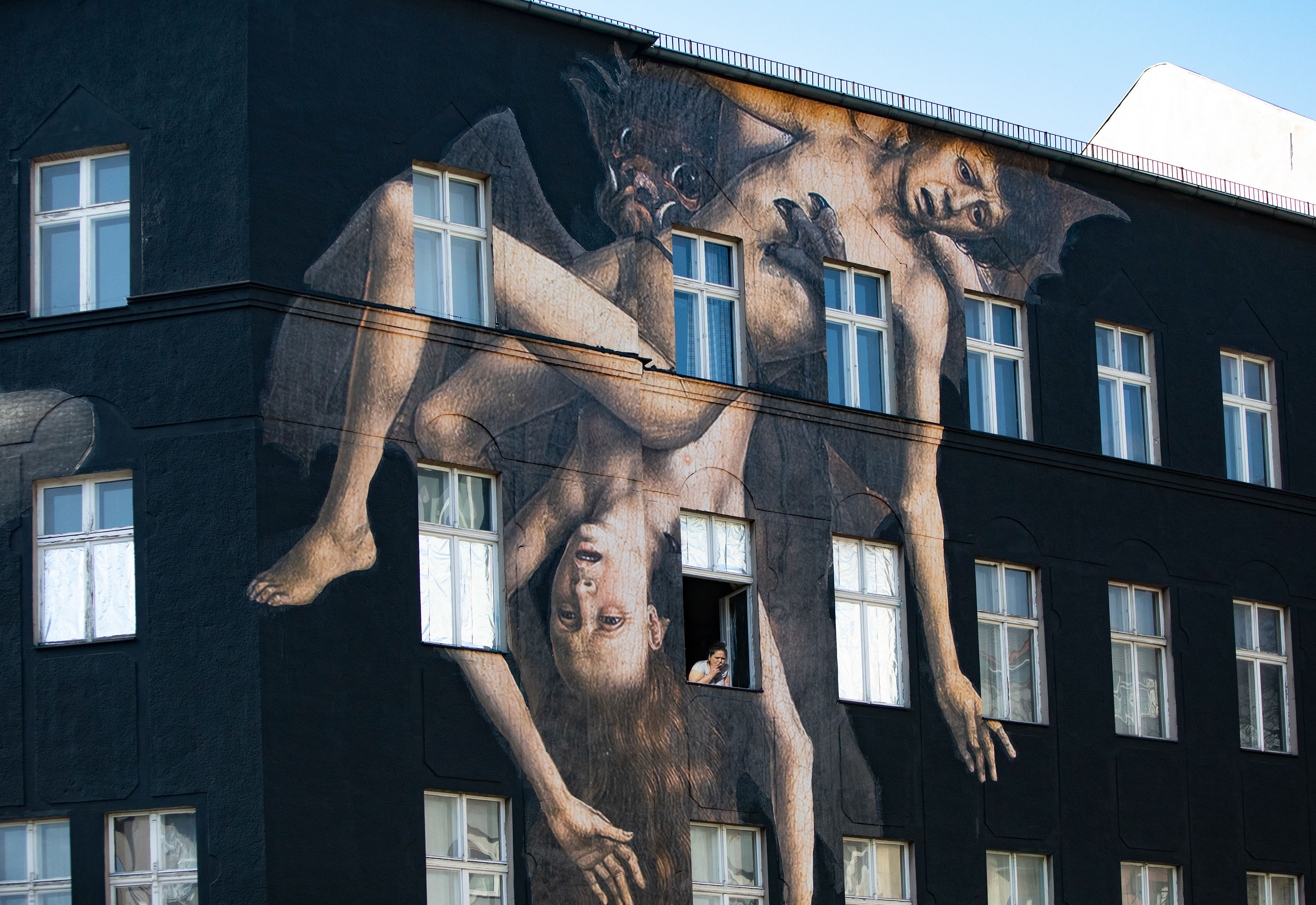 Eine schwarze Hauswand, auf der ein übergroßes Gemälde zu sehen ist – und in der Mitte schaut ein Mensch aus einem Fenster.