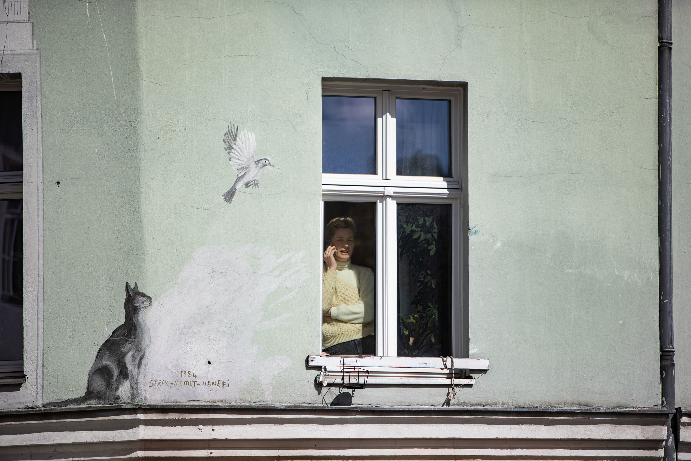 Eine Frau steht hinter einem geschlossenen Fenster und Telefoniert, die Hauswand ist verziert mit einem gemalten Vogel und einer Katze.