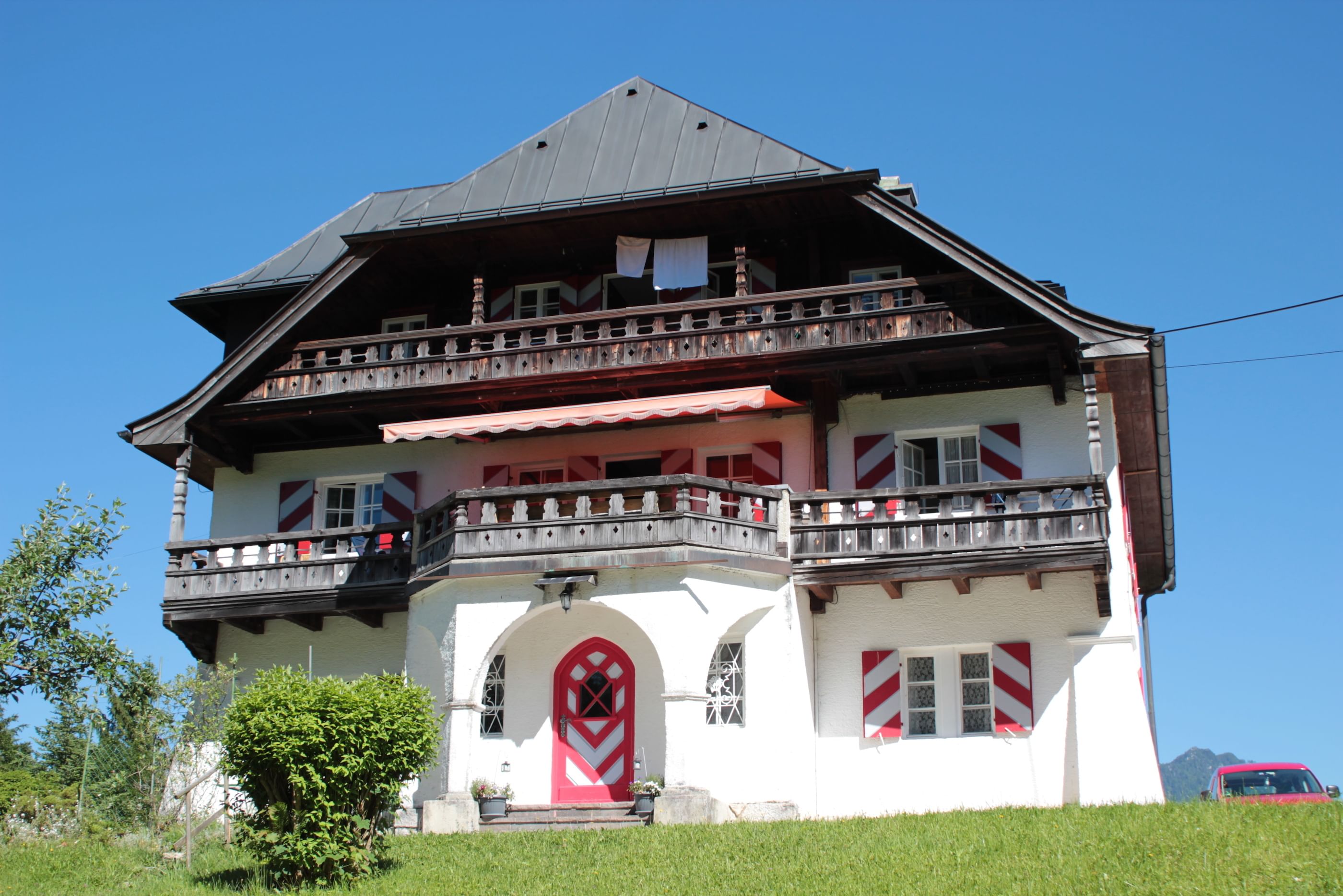 Ein altes Haus mit rotgestreiften Fensterbedeckungen steht in der Landschaft, davor eine grüne Wiese. 