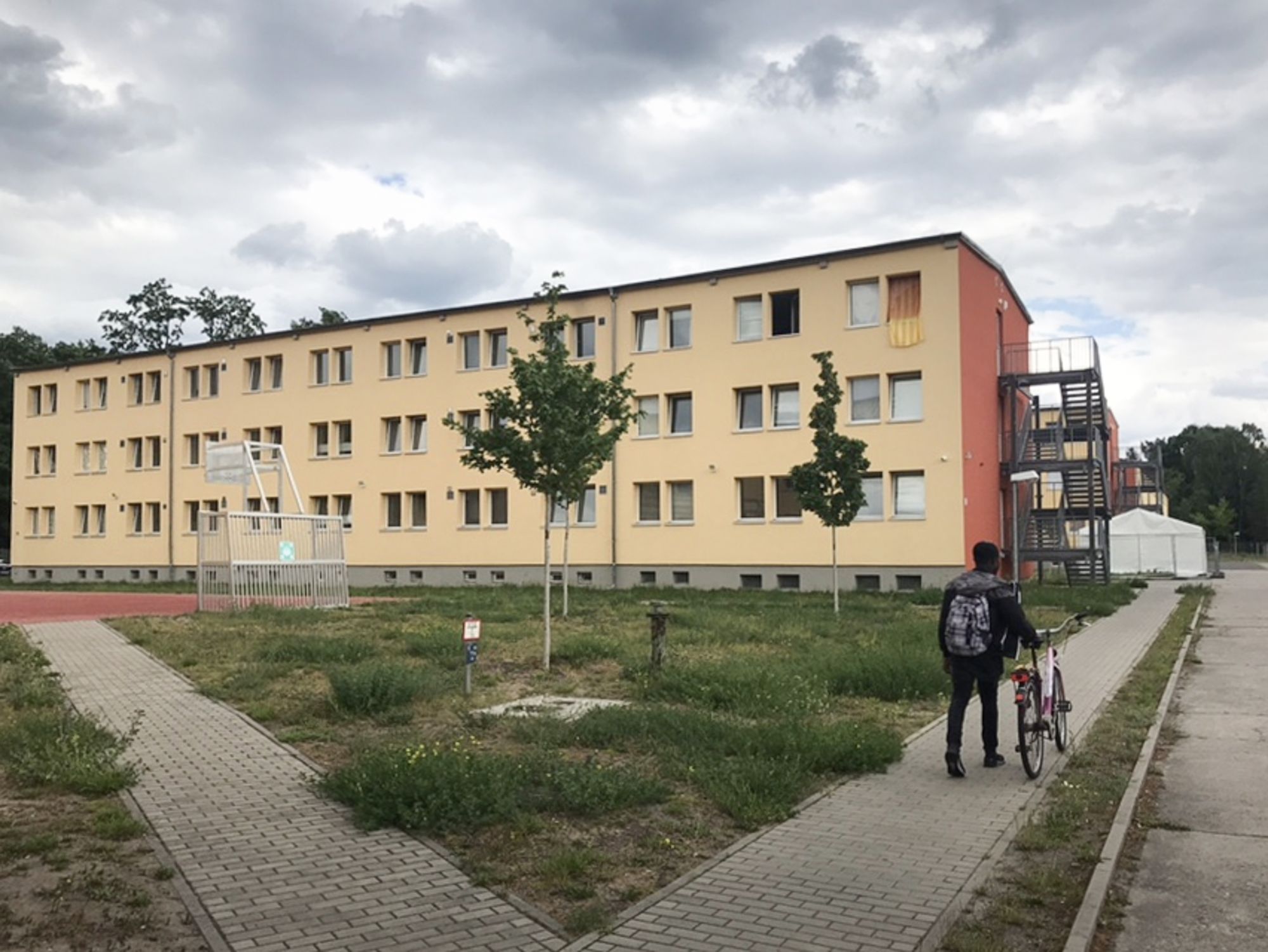 Wir sehen ein dreistöckiges Haus mit flachem Dach, davor ein Fußballplatz neben einer Wiese – Moussa schiebt auf dem Weg am Haus vorbei sein Fahrrad.