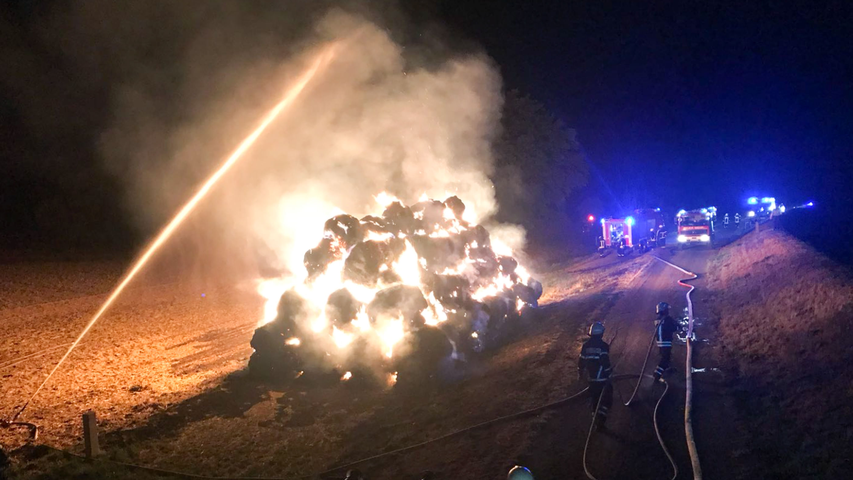 Blick aus erhöhter Position auf viele brennende Heuballen, die Feuerwehr löscht von der Seite, im Hintergrund Blaulichter und Feuerwehrfahrzeuge.