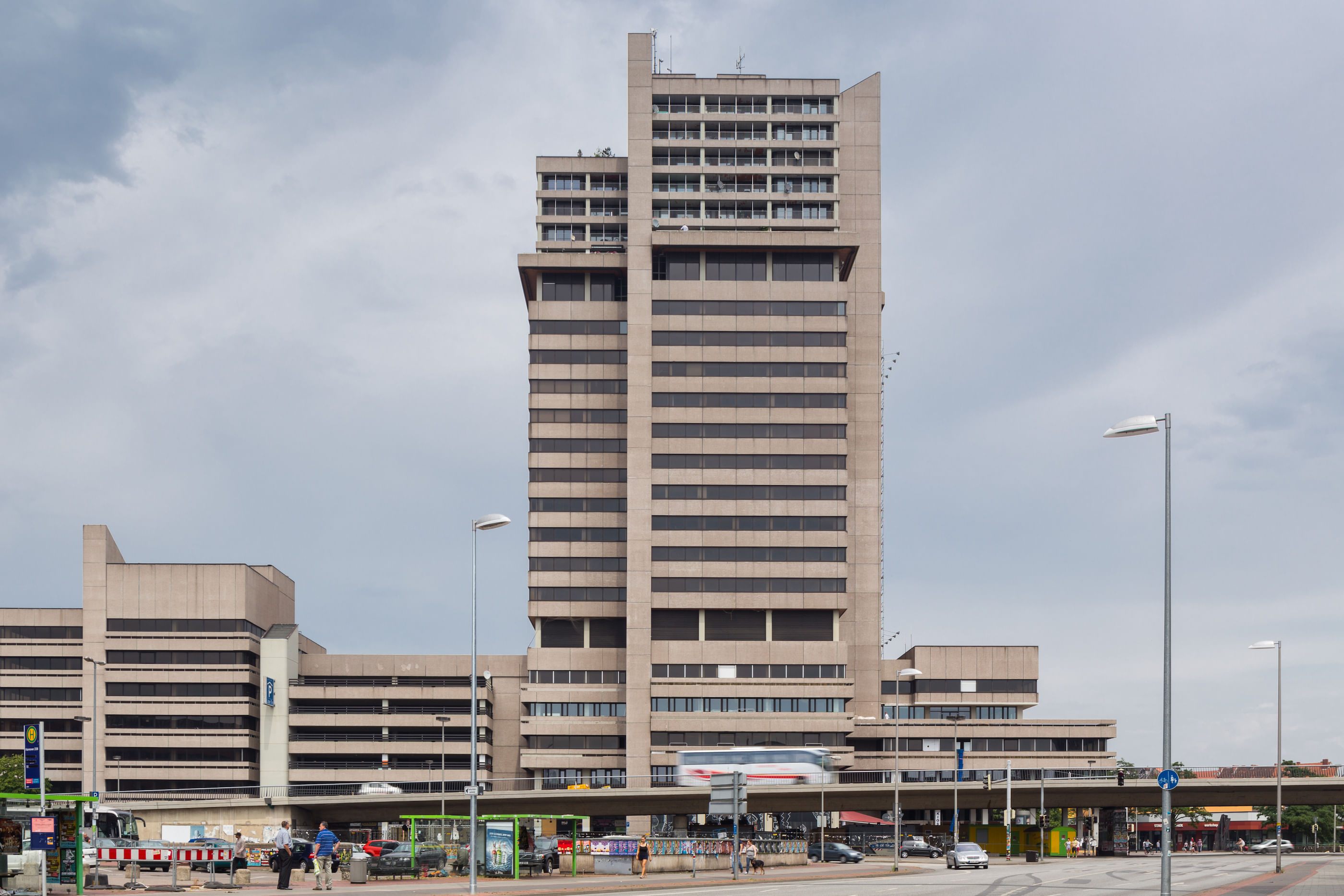 Wir sehen aus der Ferne ein Hochhaus. Es ist grau und wirkt relativ kühl. Der Himmel ist bewölkt und am unteren Bildrand sehen wir Menschen, die in Busse steigen.