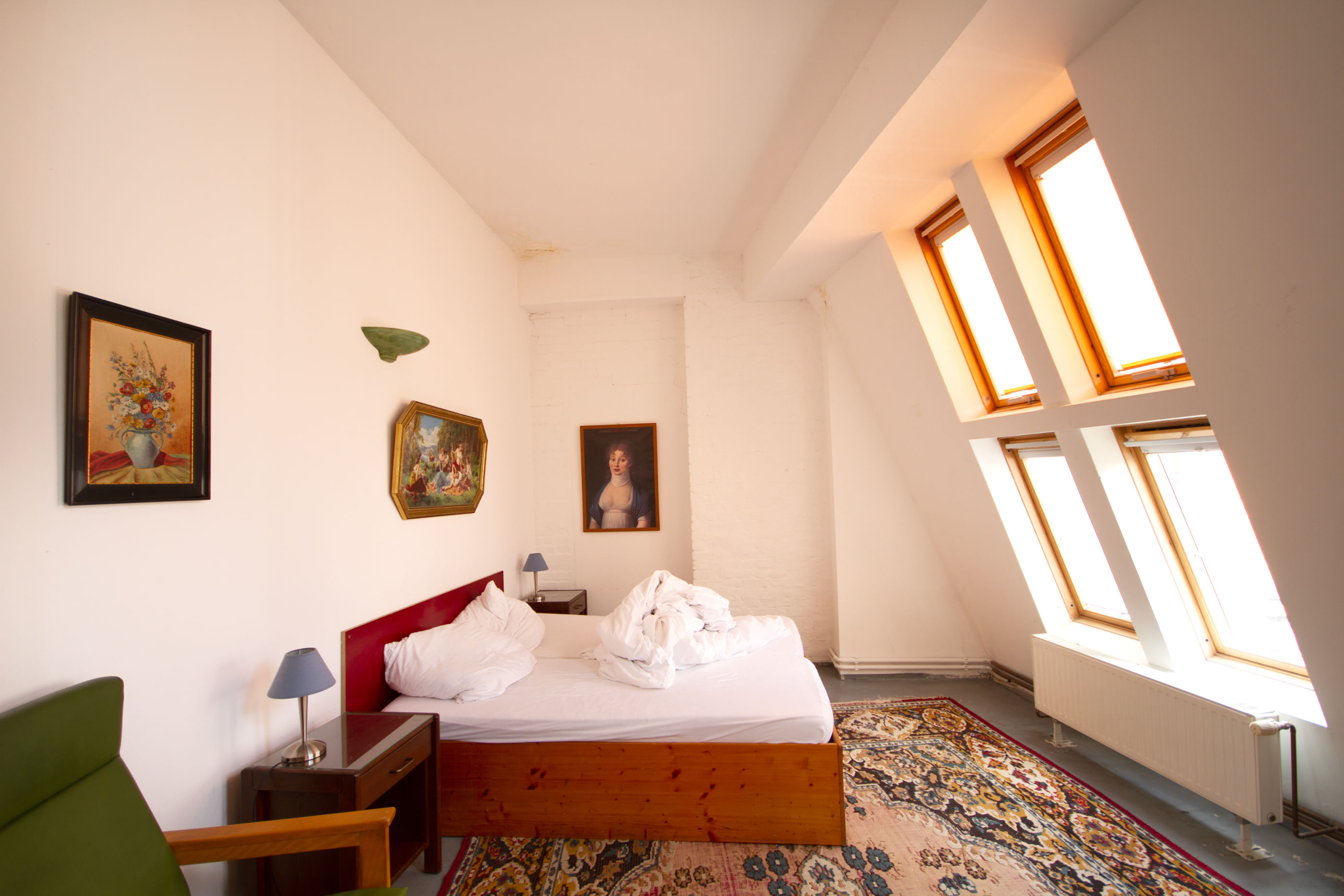 Wir sehen ein Zimmer, in dem neben Sesselstühlen ein Bett mit weißem Bezug steht – und rechts davon vier große, helle Fenster, die von außen Licht einlassen.