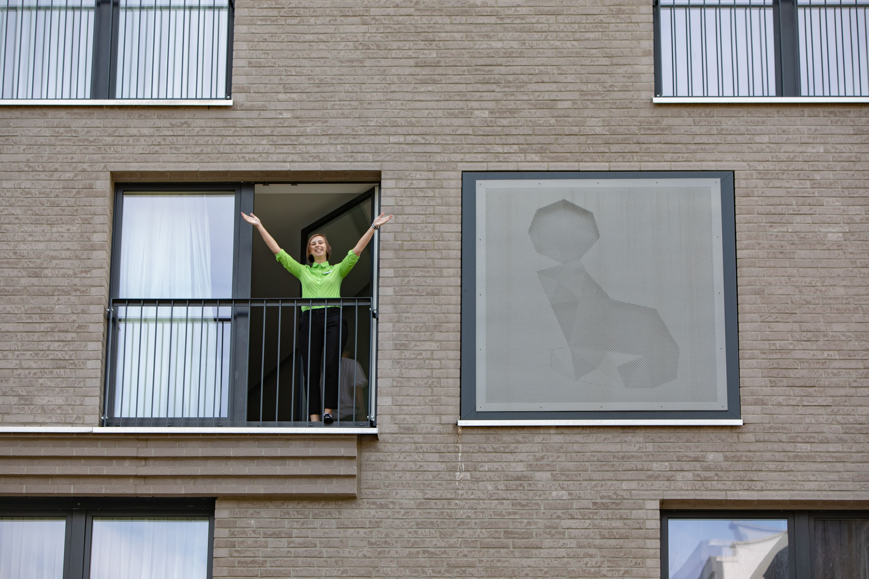 Wir sehen eine Frau, die mit offenen Armen an einem Fenster steht und uns begrüßt – sie trägt ein grünes Oberteil.