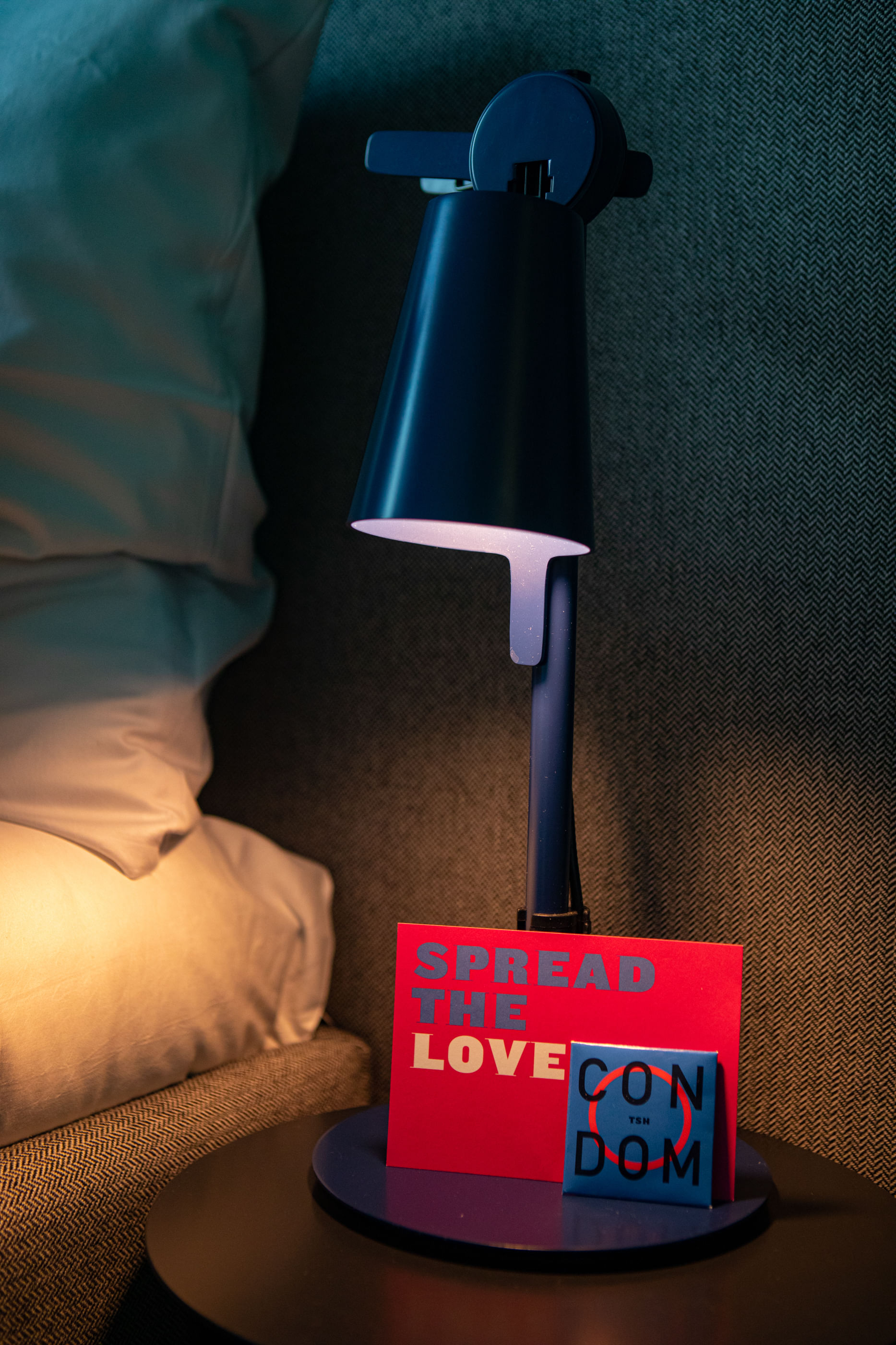 Wir sehen eine Nachttischlampe, davor ein Kondom und eine Karte mit der Aufschrift: Spread the love