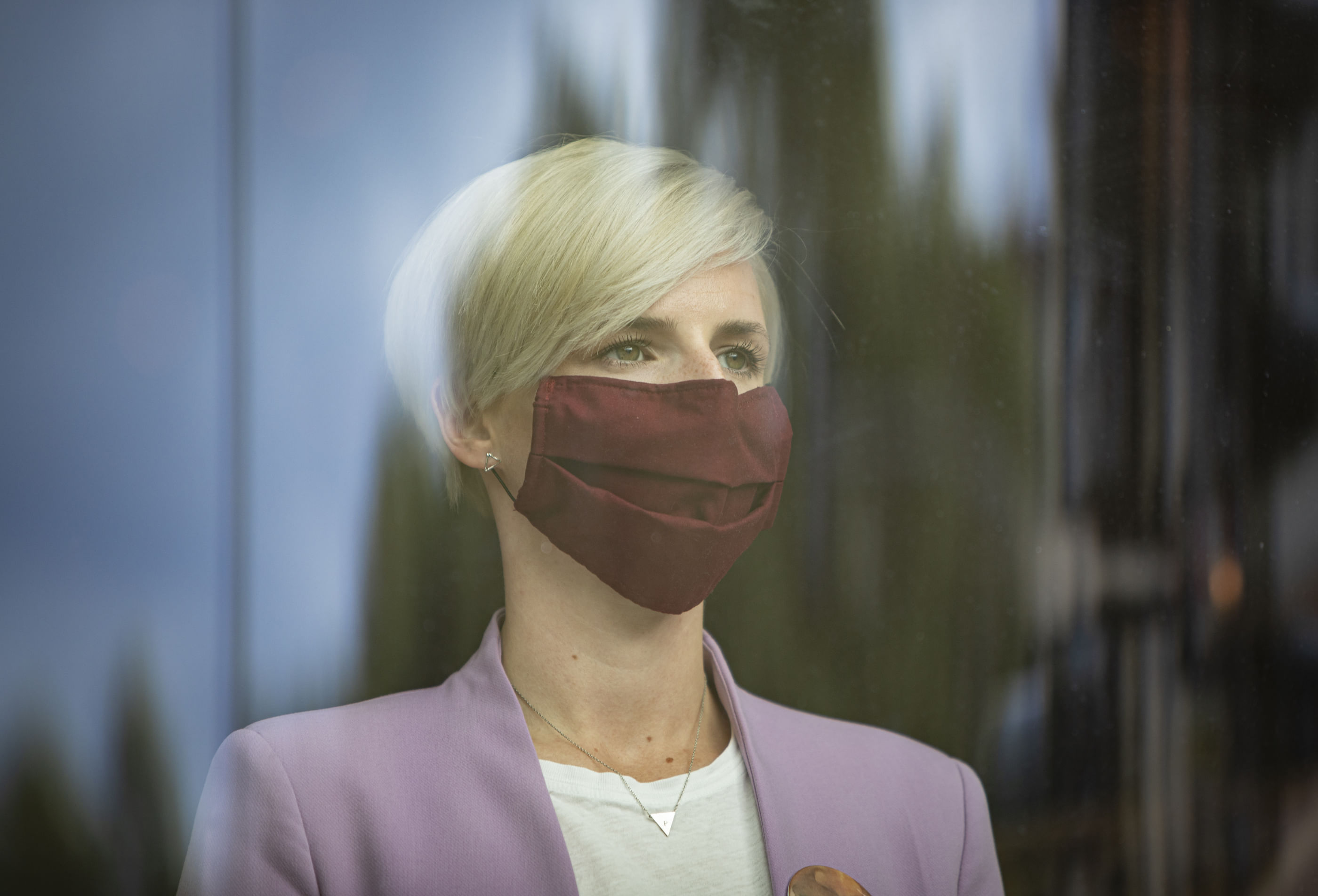 Wir sehen eine blode Frau mit Maske und lilanem Jaquette, die aus dem Fenster schaut