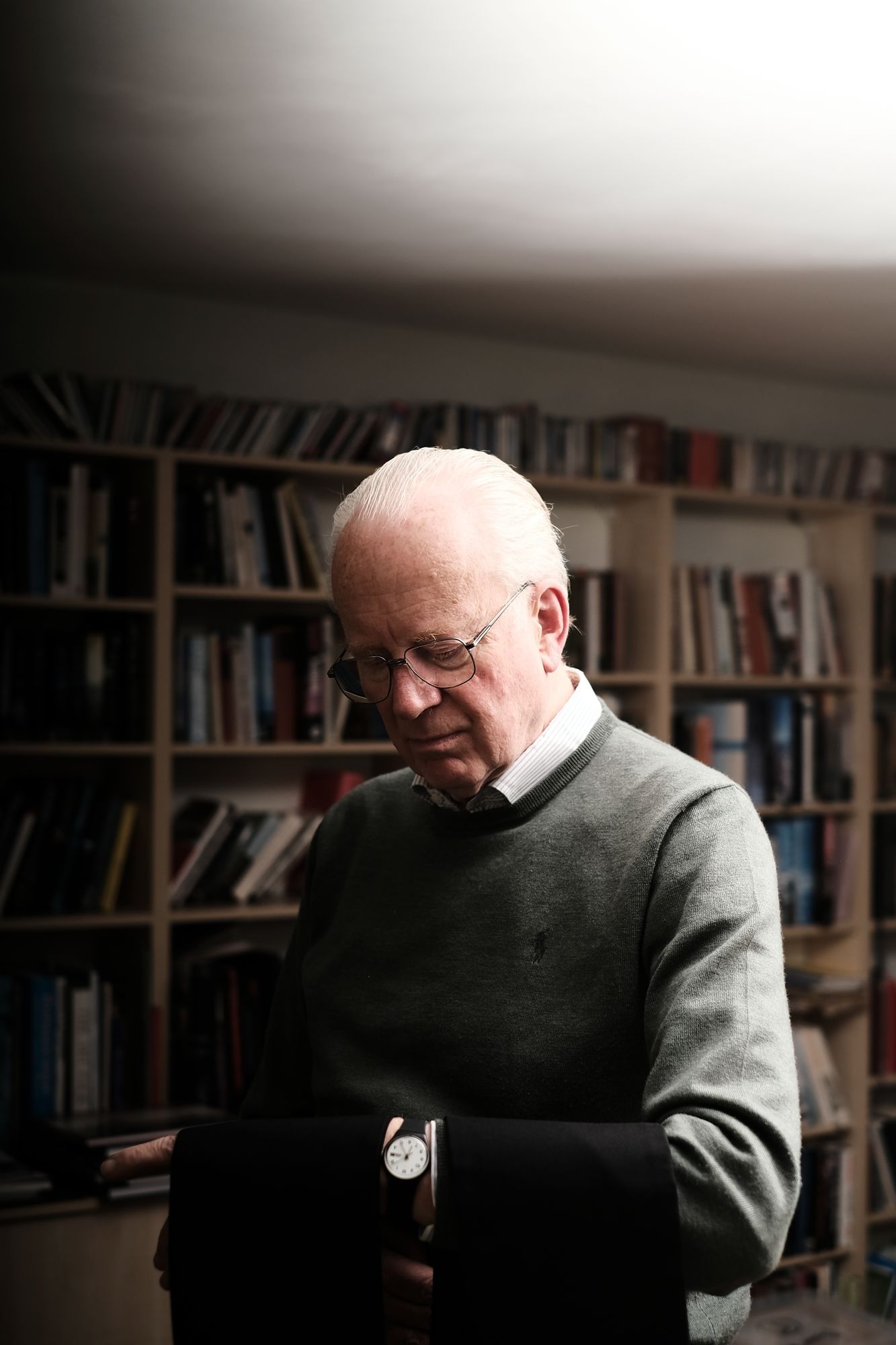 Nicholas Frayling, ein älterer Mann mit Brille und grauem Harr steht in seinem Wohnzimmer vor einem Bücherregal. Er hält den linken Arm hoch, über dem schwarze Tücher liegen.