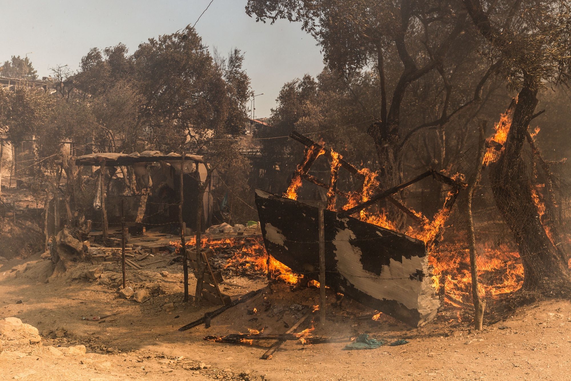 Wir sehen abbrennende und abgebrannte Zeltstrukturen hinter einem noch stehenden Zaun zwischen ein paar Bäumen, die ebenfalls Feuer fangen. Ein trostloses Bild.