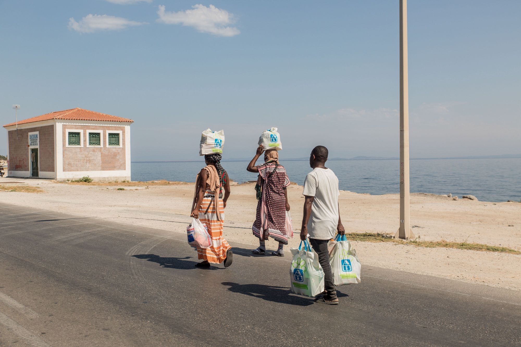 Wir sehen drei Personen, die mit vollen Einkaufstüten eine Straße entlanggehen, im Hintergrund das Meer. Zwei Menschen tragen zusätzlich vollgepackte Tüten auf dem Kopf.