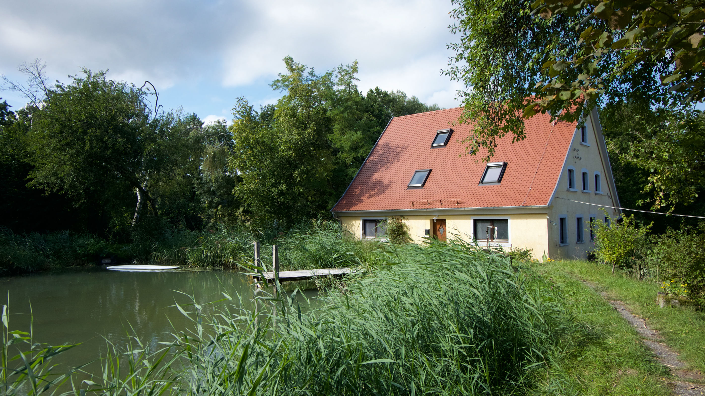 Blick auf ein Haus, gelegen an einem kleinen Teich. Im Vordergrund wilde Gräser und ein kleiner Weg.