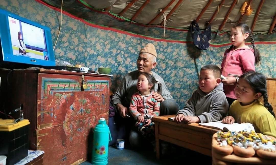 Eine Gruppe Kinder sitzt mit einem älteren männlich gelesenen Familienmitglied in einer Jurte vor dem Fernseher in welchem Unterricht läuft.
