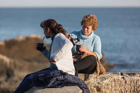 Zwei weiblich gelesen Personen trinken Mate Tee auf einer Mauer an der Atlantikküste