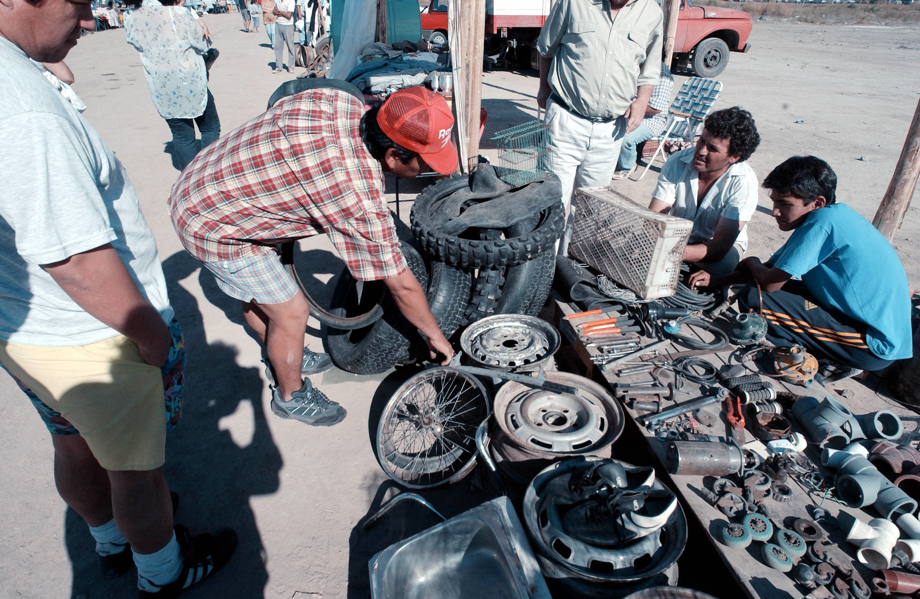 Eine Gruppe von vier Männern steht auf den Seiten einer adhoc Auslage welche mit unterschiedlichen, vorrangig Automobil, Ersatz- und Einzel-teilen beladen ist.