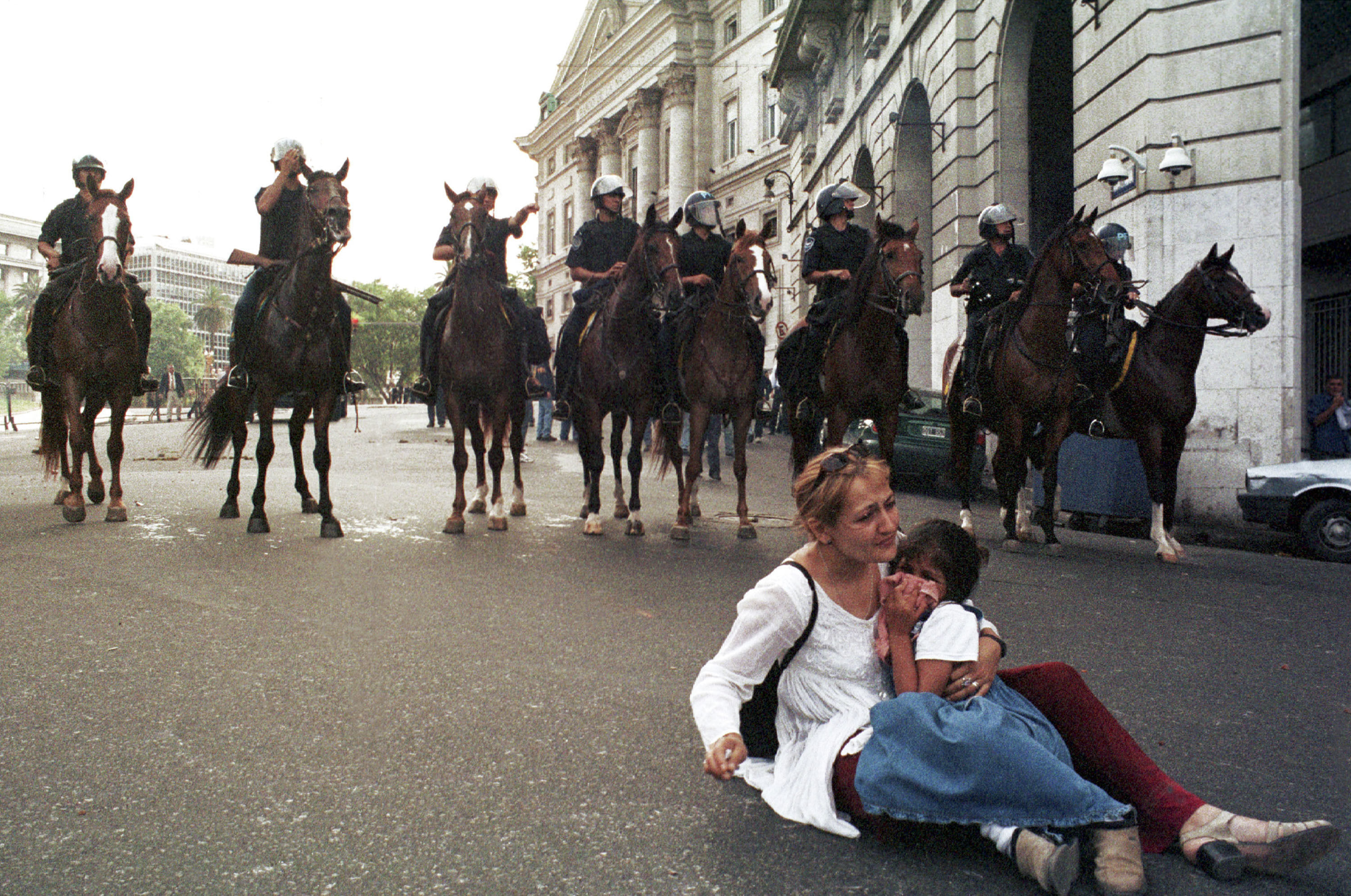 Eine Frau sitzt mit einem Kind, welches sichtlich verängstigt ist, mit dem Rücken zu einer Postenkette von berittener Polizist:innen