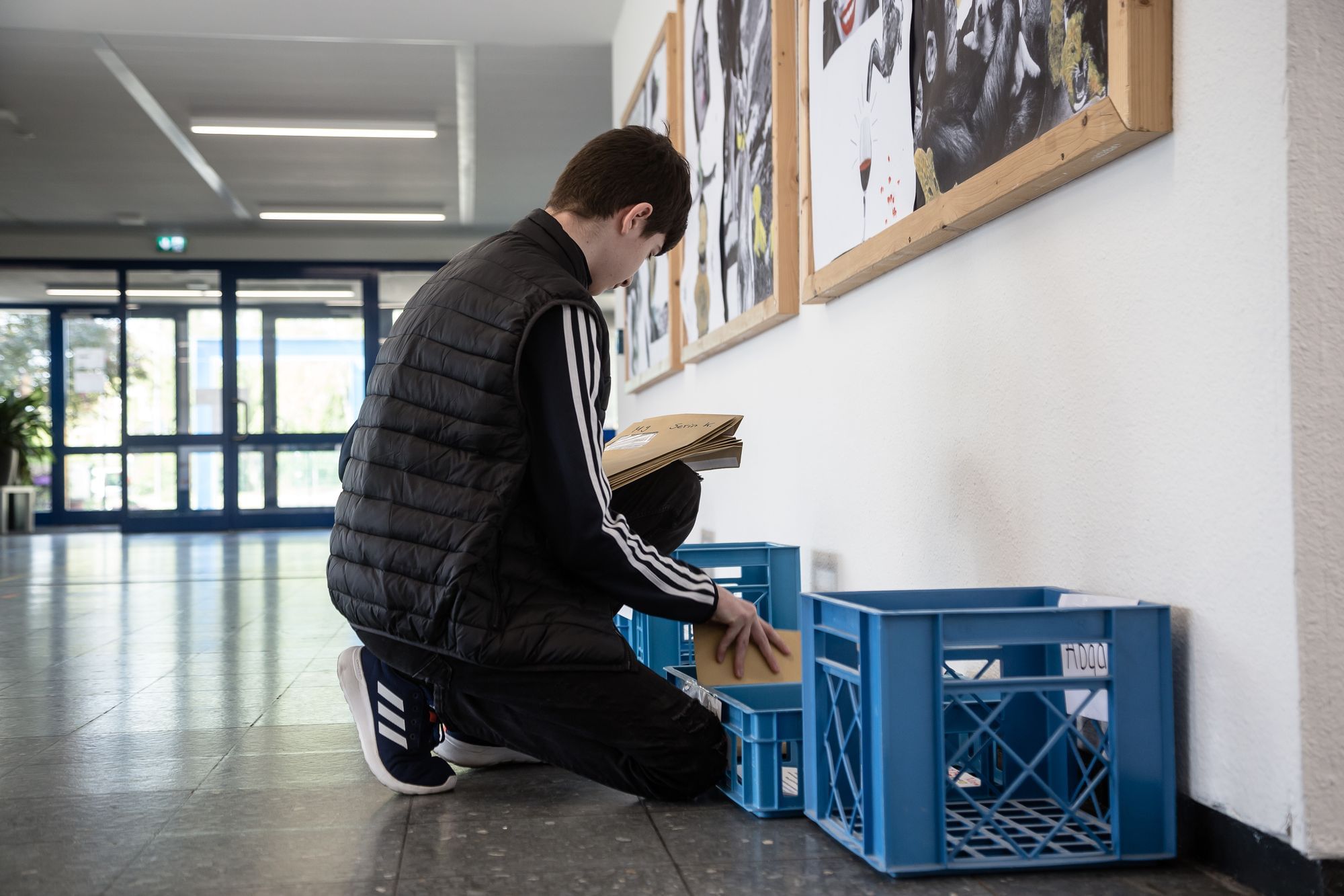 Ein Schüler hält mehrere Briefumschläge in der während er vor Plastikablagekörben kniet.