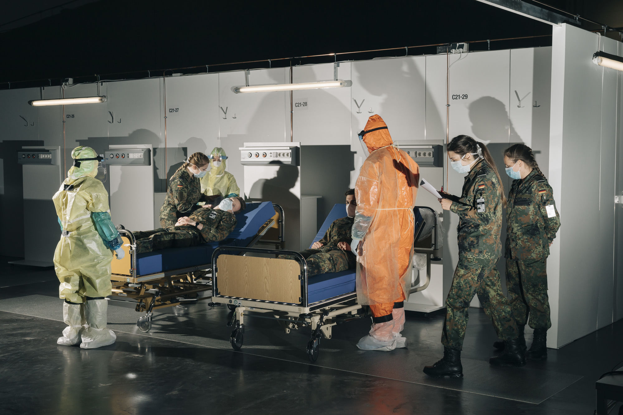 Junge Soldat:innen bei einer Übung im Behelfskrankenhaus Hannover. Zwei von ihnen liegen auf Betten und spielen Covid-Patient:innen, die anderen tragen Schutzanzug oder Bundeswehr-Uniform.