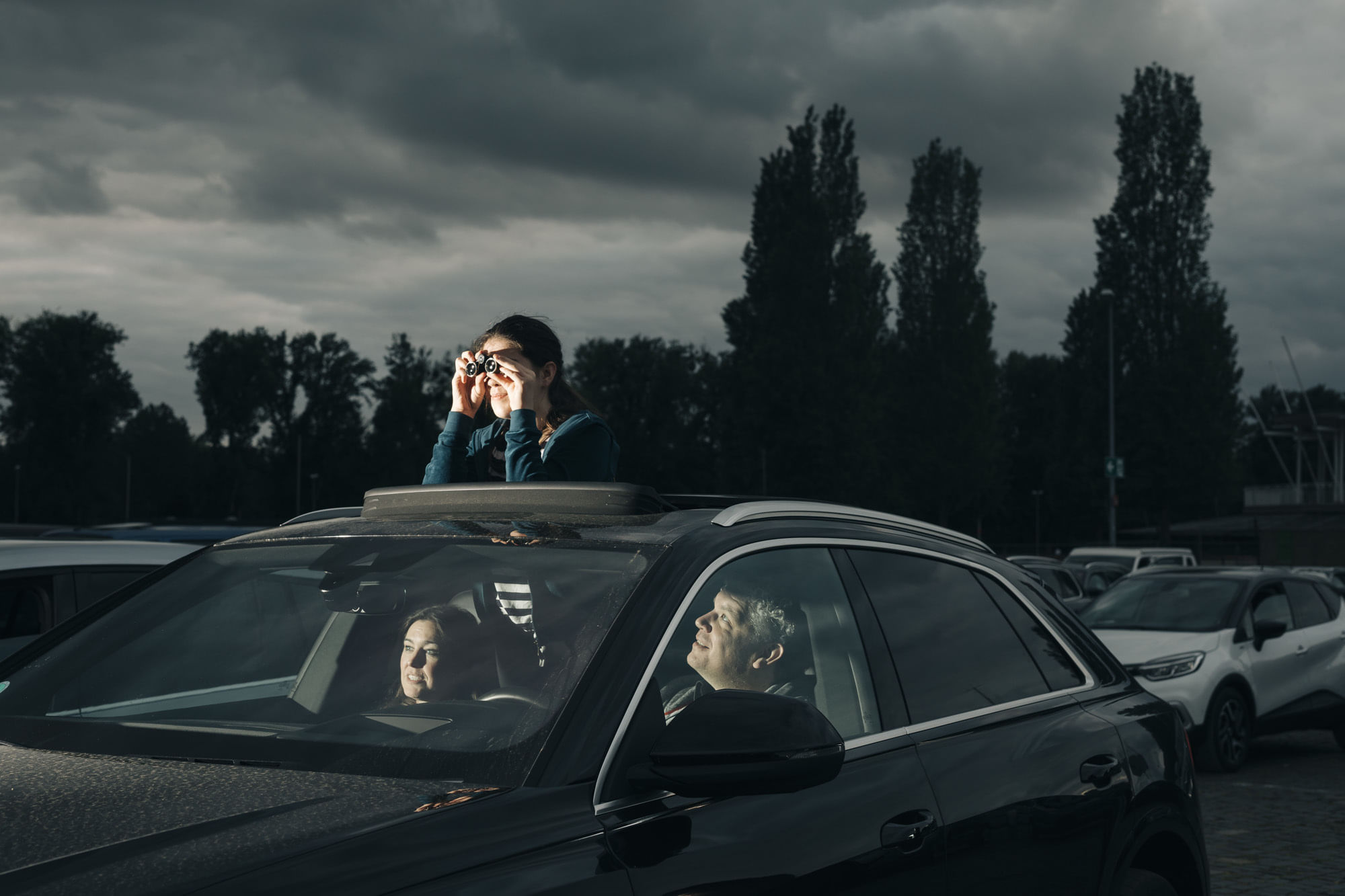 Eine Familie sitzt in ihrem Auto. Die Tochter guckt mit einem Opernglas in der Hand aus dem Dachfenster.