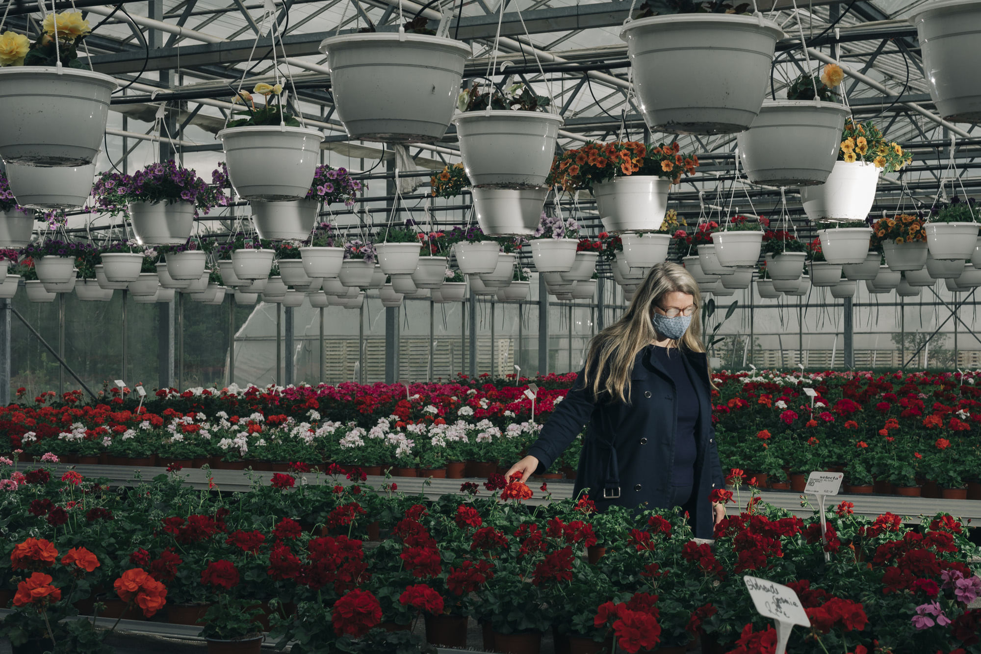 Eine Frau läuft durch einen Blumenladen. Sie trägt Maske, ihre Brille ist beschlagen. Sie berührt im Vorbeigehen eine Blume.