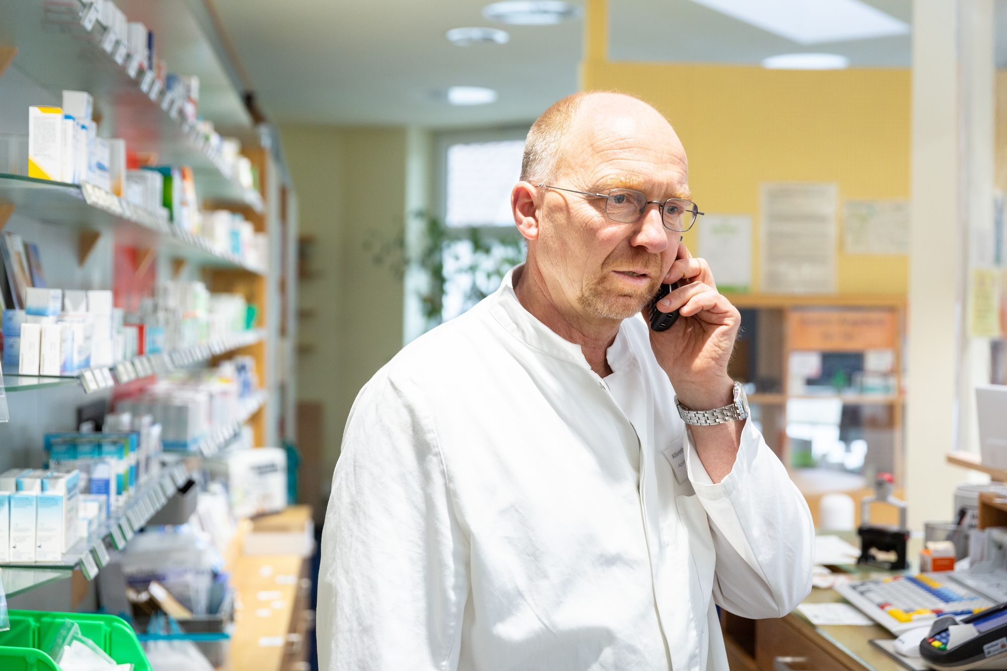 Hier sieht man, wie ein Mann in einem weißen Kittel (ein Apotheker) in seinem Geschäft telefoniert.