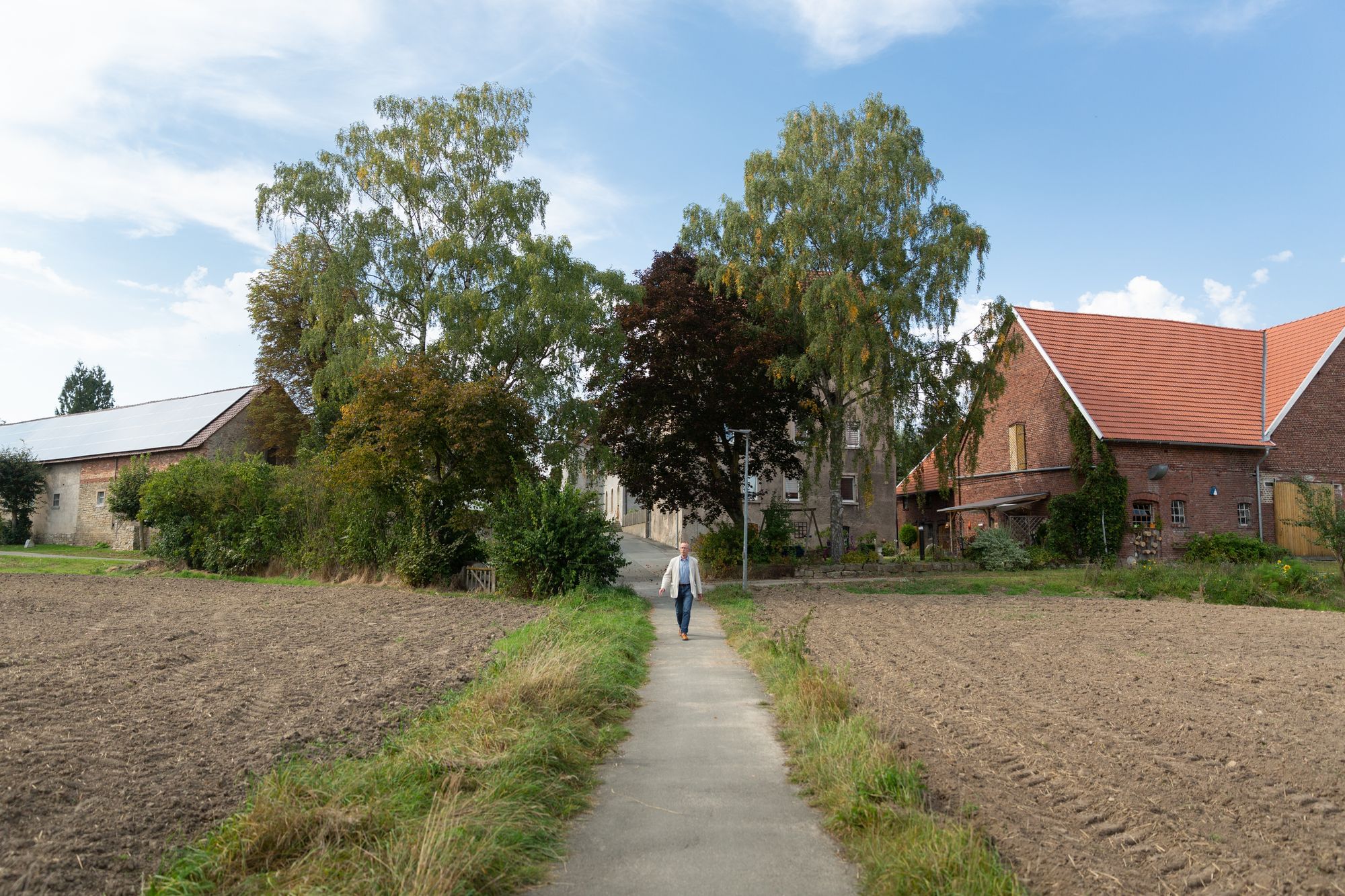 En Mann läuft auf einer Straße zwischen zwei Feldern, links und rechts des Weges ein großes Haus.