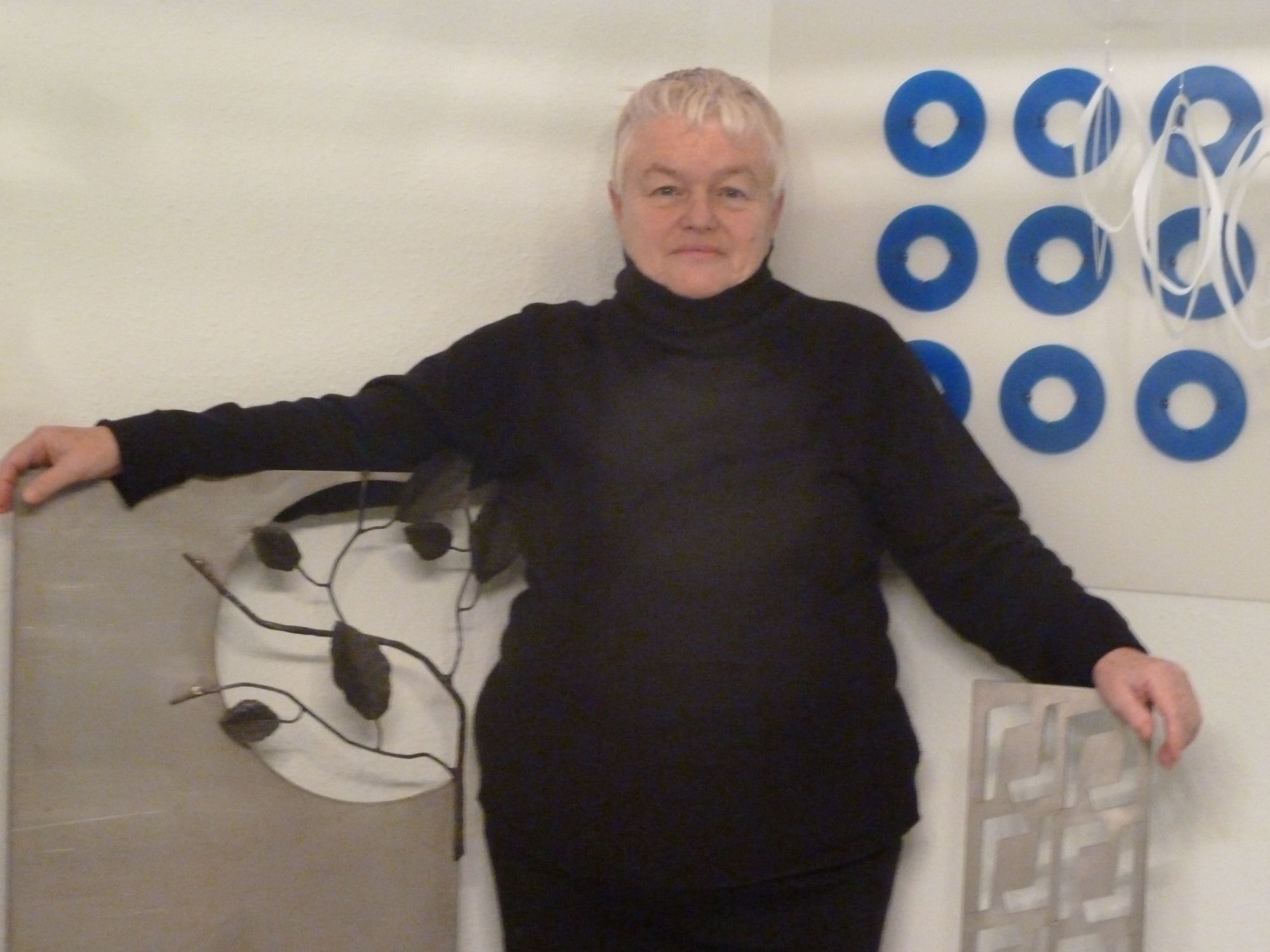 Eine Frau mit weißem, kurzem Haar steht zwischen von ihr geschaffenen moderne Objektkunstwerken.