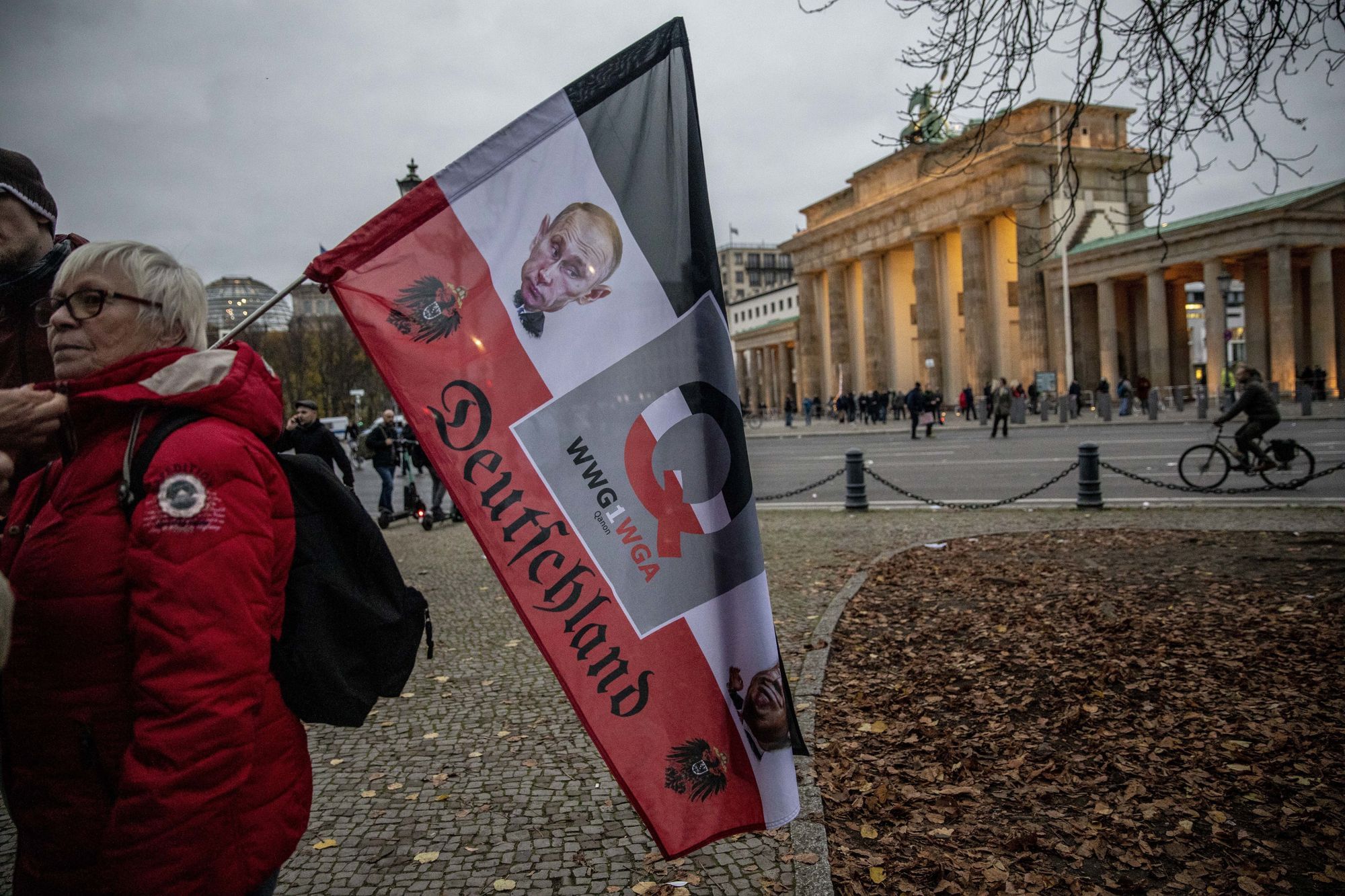 Eine Frau steht mit einer Flagge des Deutschen (Kaiser-)Reichs mit Emblemen und Chiffren der Q-Verschwörung vor dem Brandenburger Tor.