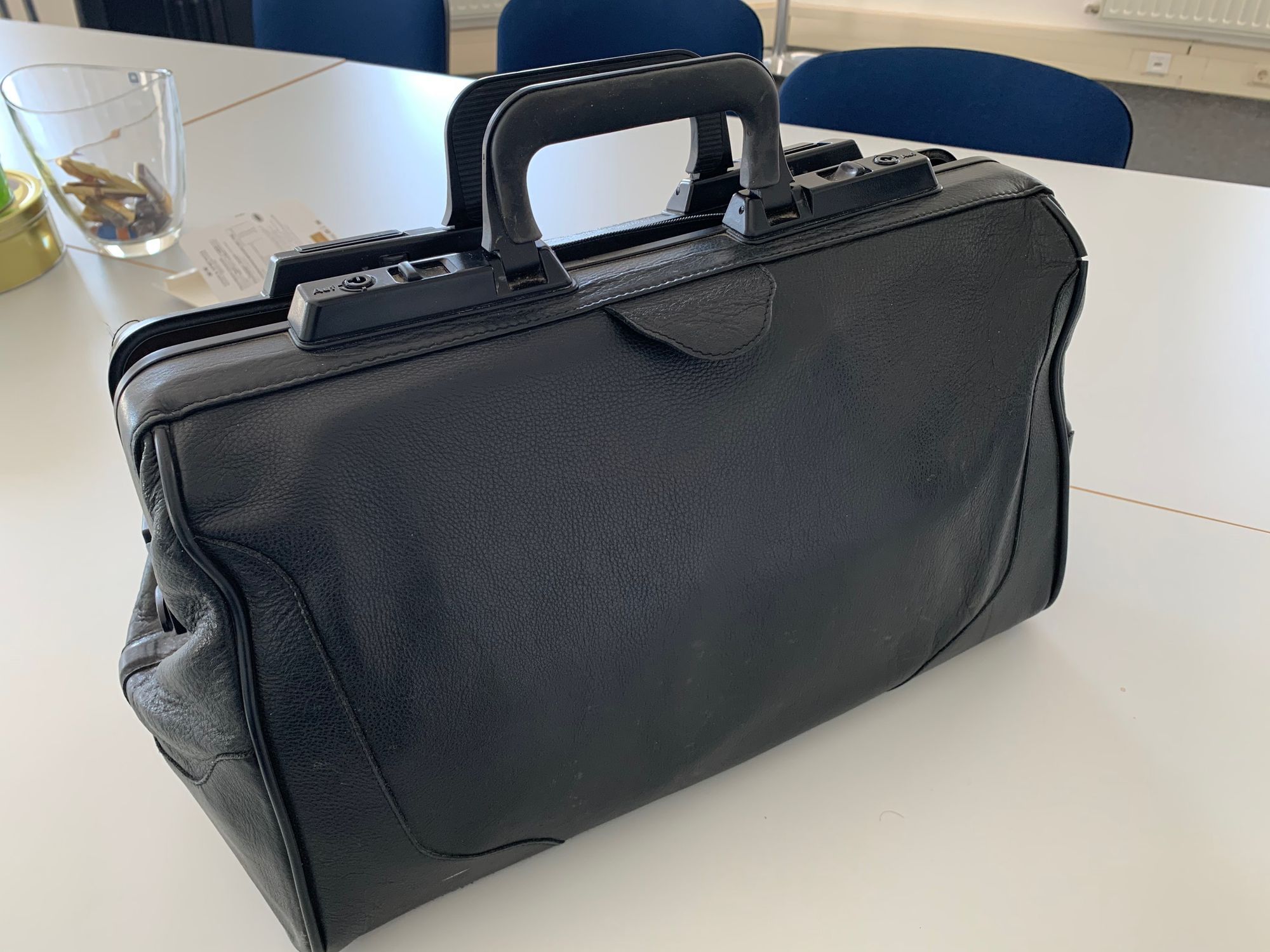 Das Bild zeigt eine schwarze Arzt-Tasche aus Leder, die auf einem Tisch steht.