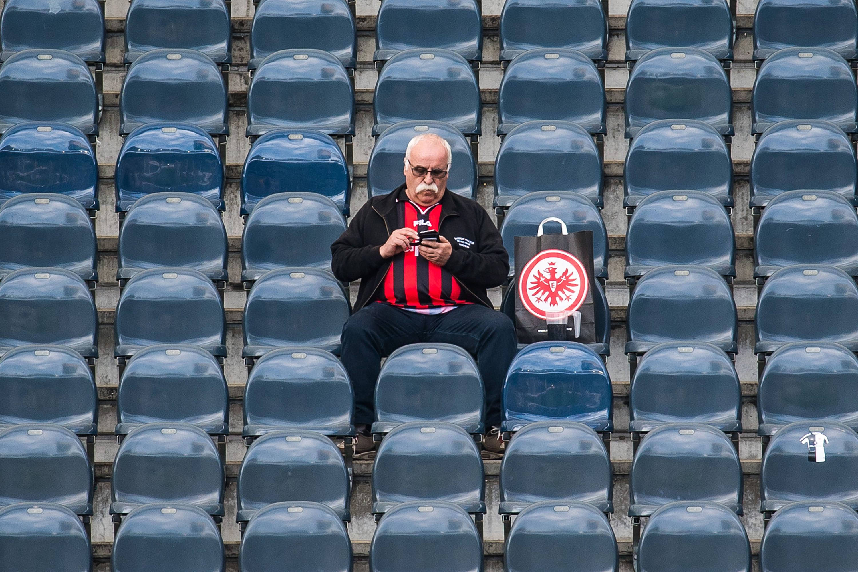 Eine männliche lesende Person ca im Alter von 50-60 Jahren, sitzt alleine auf der Tribüne und schaut auf ihr Smartphone.