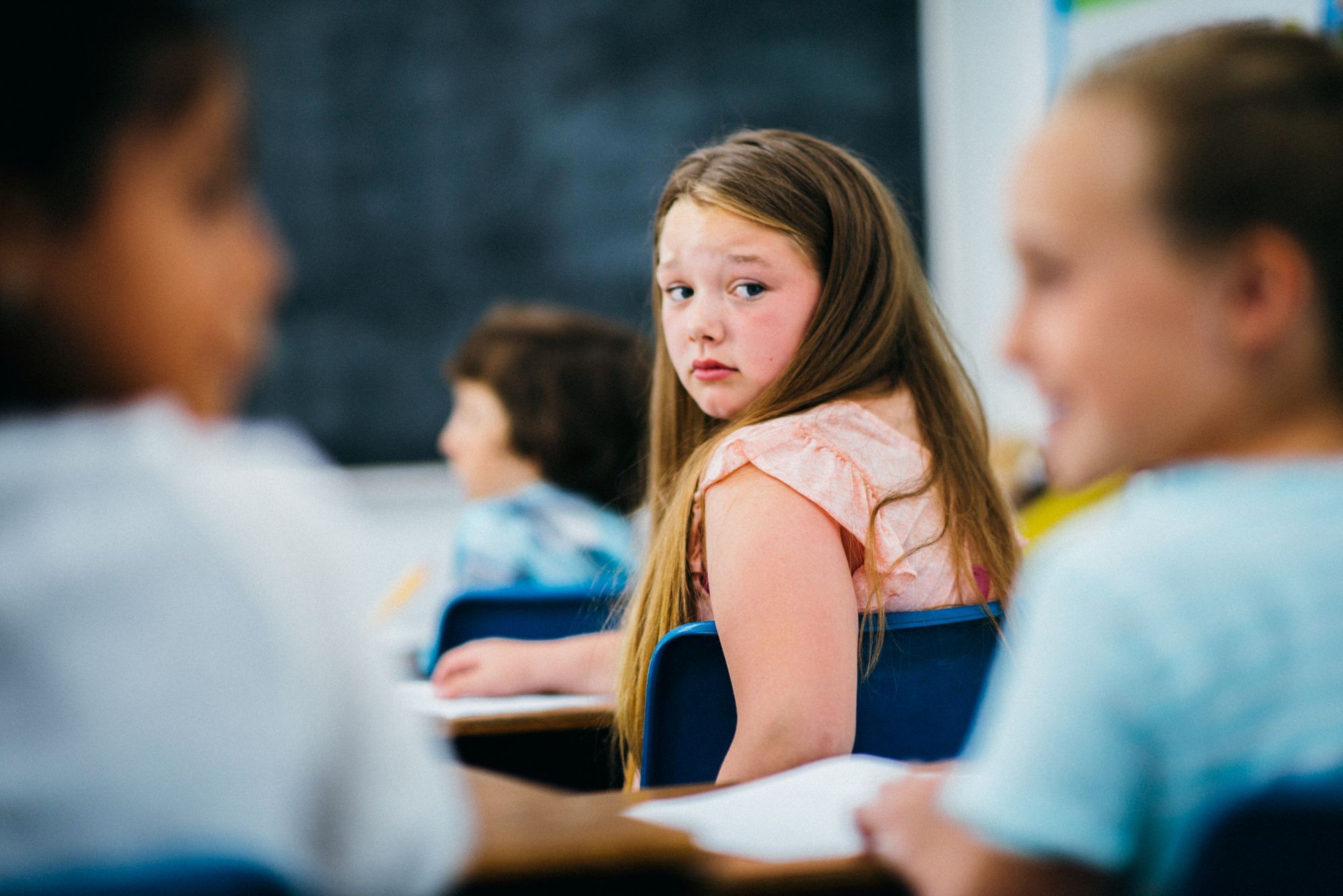Eine traurige wirkende Schülerin, welche sich einsam zu fühlen scheint, blickt im Klassenzimmer nach hinten zu den dort sitzenden Kindern.