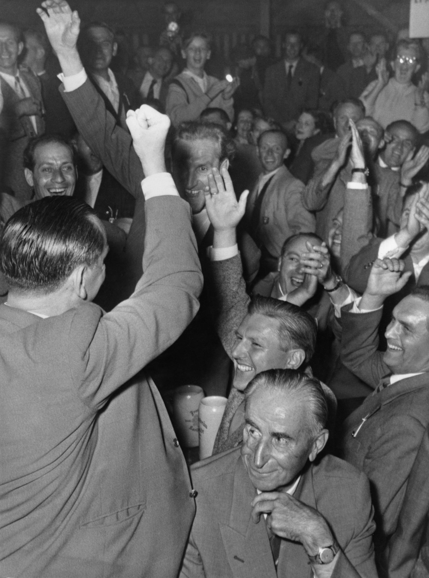 Schwarz-weiß Bild, dicht and dicht stehen ausgelassene Männer in den grausten Anzügen zusammen. Einer der Männer steht und den Rück zugewandt und die rechte Faust erhoben die anderen ansporenend. Einer der Männer sieht glücklich funkeln begeisternd aus.
