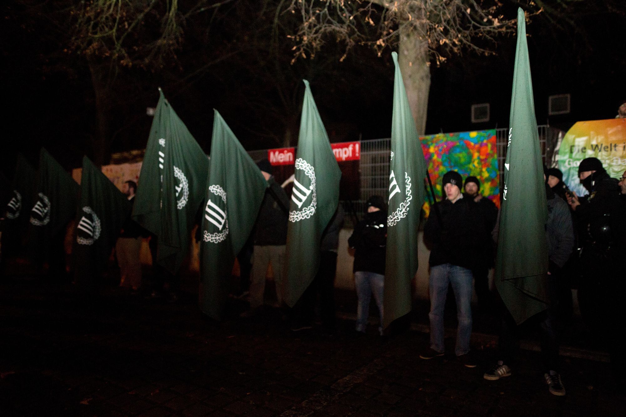 Geblitzte Nachtaufnahme eines in Reihe stehenden Fahnenspaliers. Die Fahnen sind grün und mit einem Lorbeerkranz welcher eine römische Drei umschließt.