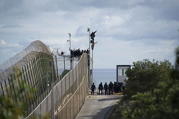 Eine Gruppe Migrant*innen überquert den Grenzzaun bei Mellila. Wir sehen den breiten Zaun und eine Person hält ihre Handhoch auf dem Zaun hängend.