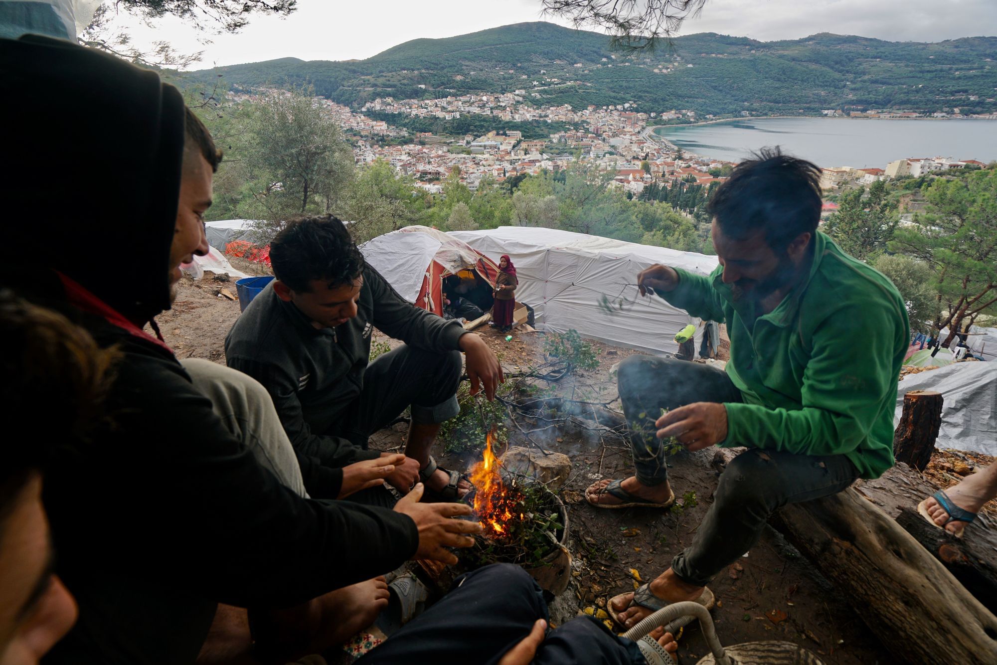 Eine Gruppe geflüchteter Menschen aus Syrien sitzt um ein Lagerfeuer, im Hintergrund sehen wir eine Bucht mit Stadt.