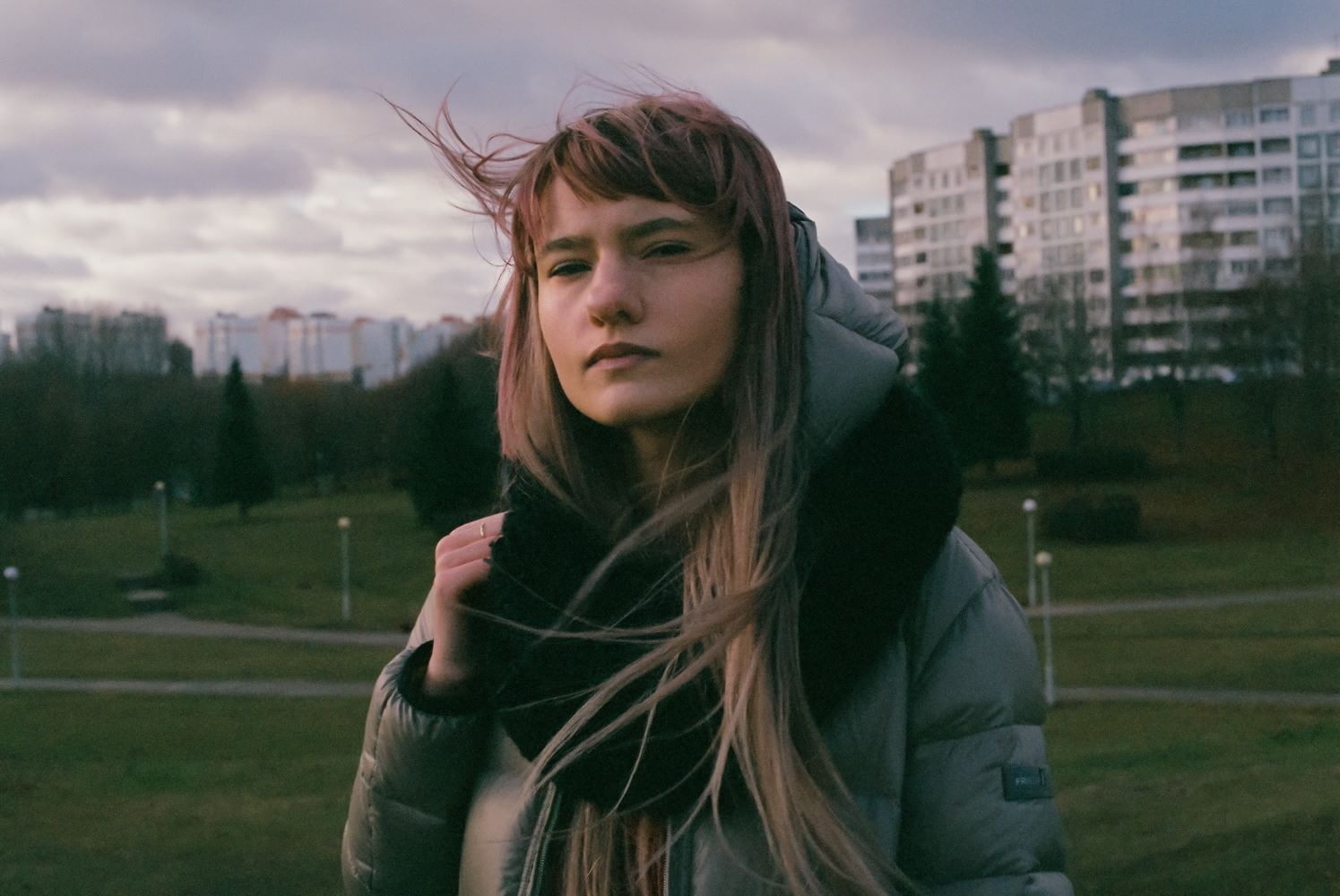 Portät einer jungen Frau vor einem Park und im Hintergrund Plattenbauten. Sie schaut entschlossen in die Kamera, ihre Augen scheinen leer.