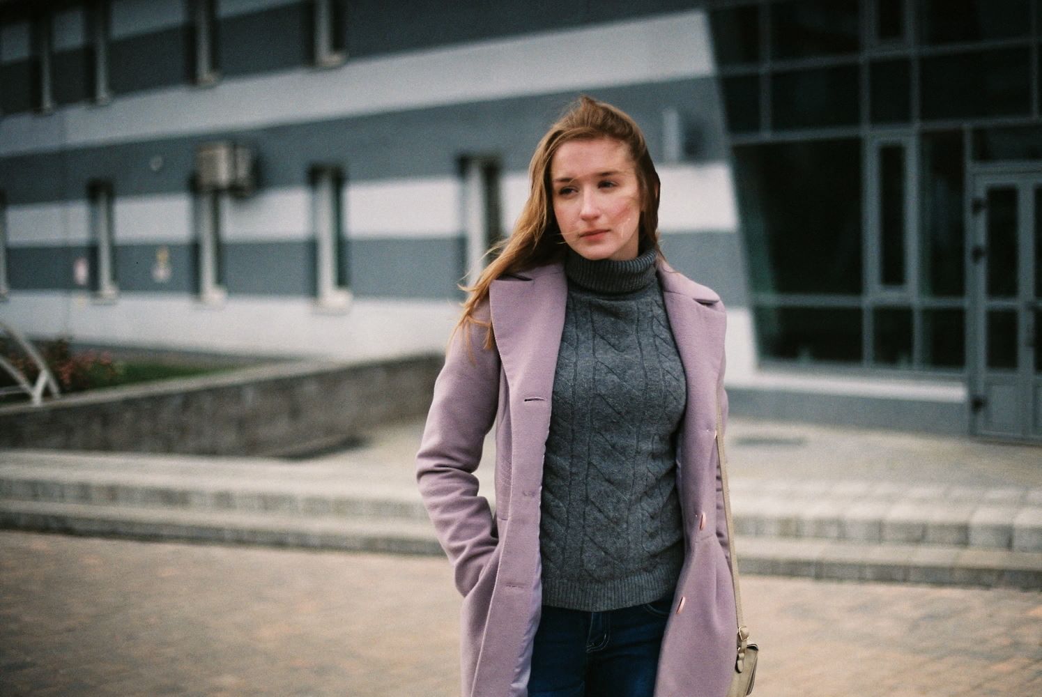 Halbkörper Porträt einer Frau vor einer Plattenbau- Wohnsiedlung. Ihr Kopf ist leicht geneigt und sie scheint nachdenklich zu sein.