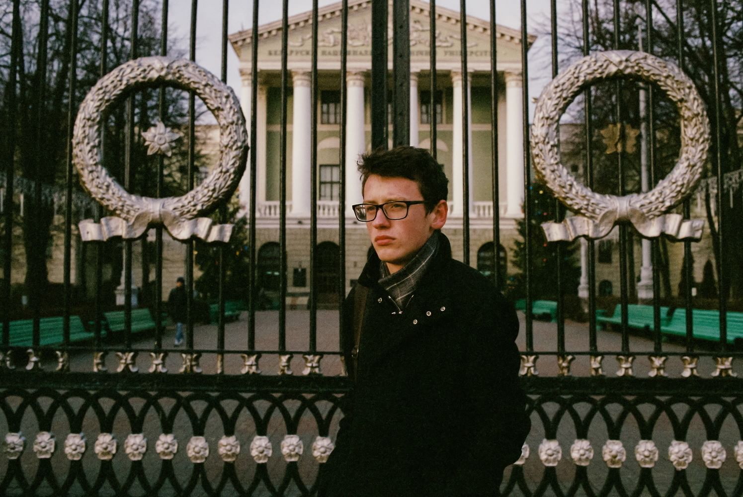 Ein junger Mann mit Brille steht vor einem reichverzierten Tor hinter welchem ein an Hellenistische Archtiktur angelehnter Platz zu sehen ist. Der Mann blick Melancholisch an der Kamera vorbei.