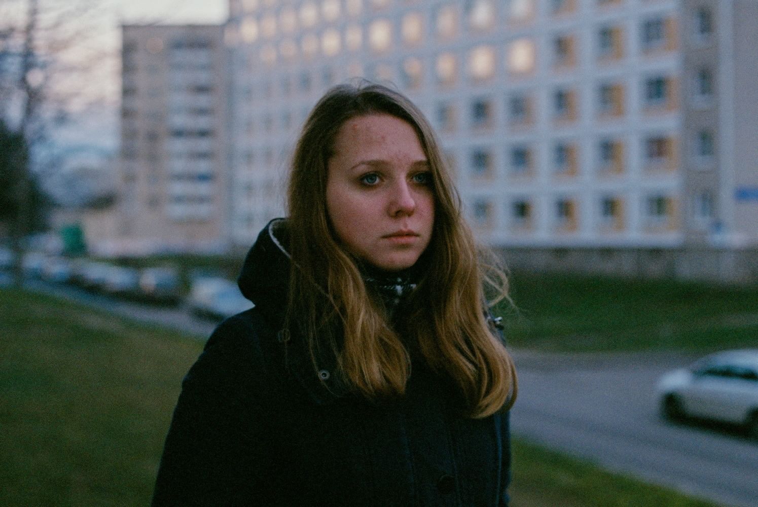 Eine junge Frau mit neutralem Gesichtsausdruck vor einer Plattenbausiedlung. Ihr Blick ist im besten Falle als Fragend zu bezeichnen.